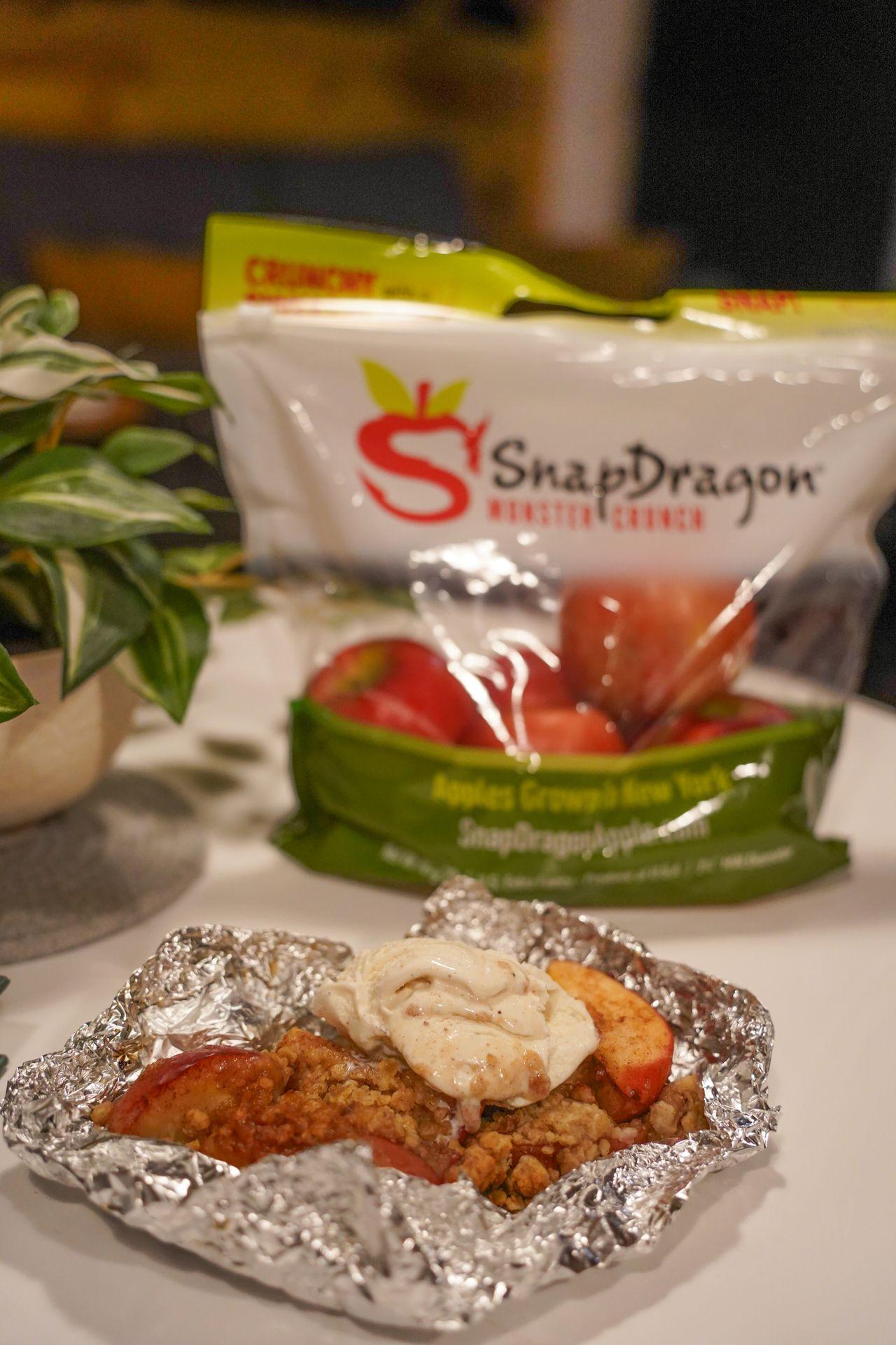 An apple tart with inside a foil packet, topped with ice cream. A bag of SnapDragon apples sits on the table in the background.