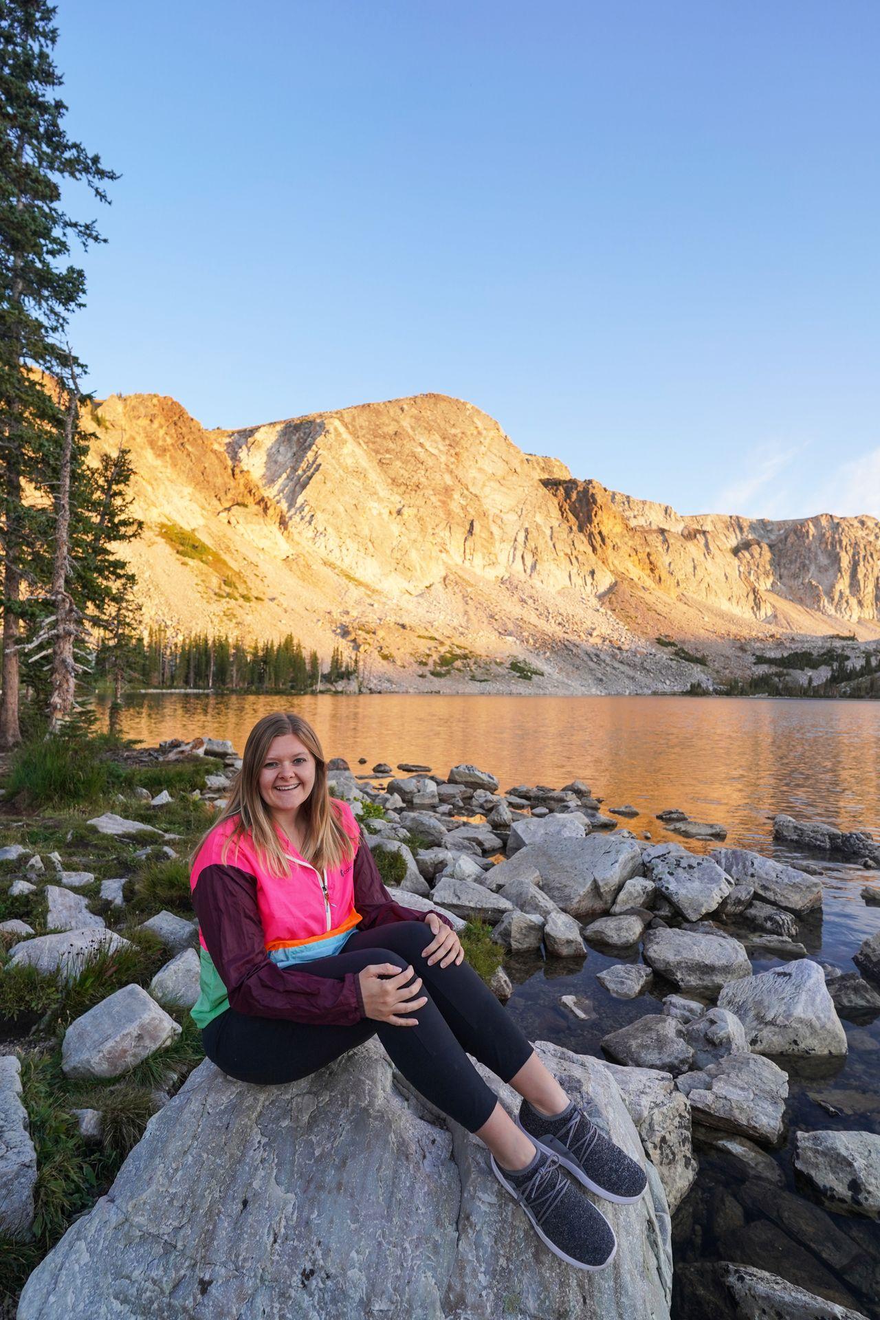 Lydia sitting on a rock next to a lake while wearing a brightly color blocked windbreaker