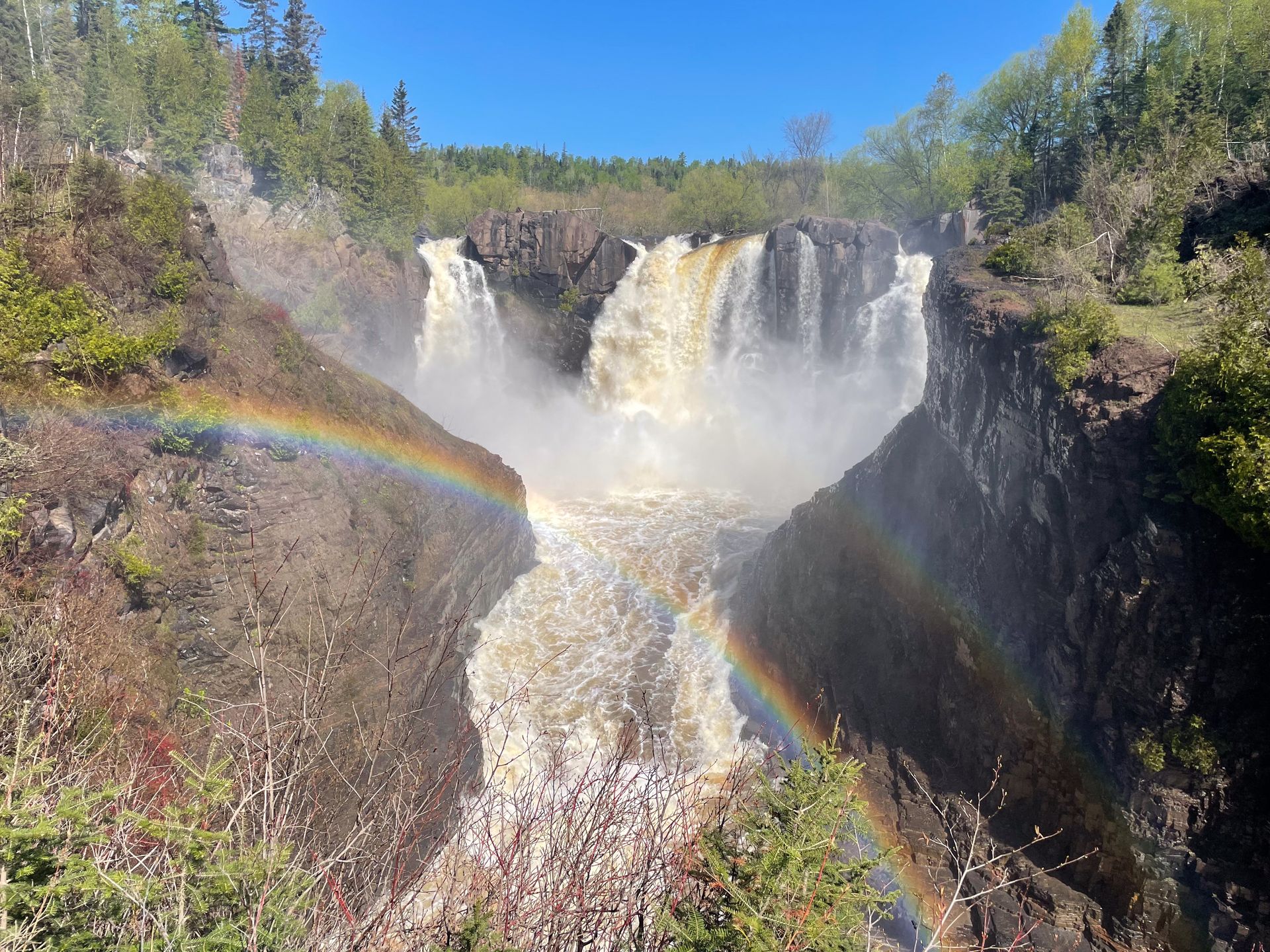 A waterfall flowing down into a black gorge from 3 directions. A rainbow is in the foreground.