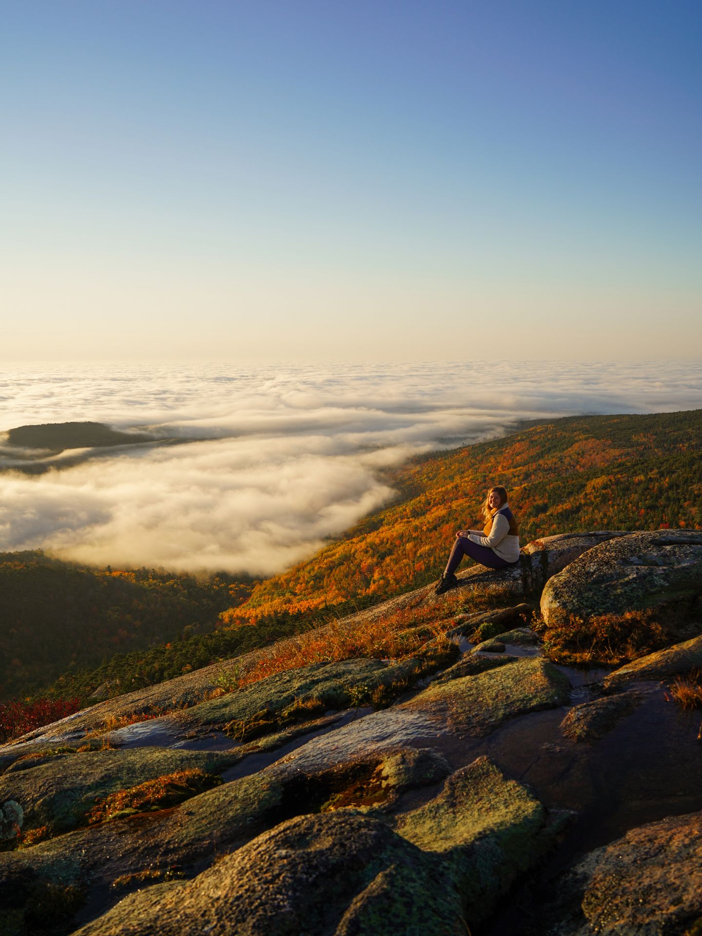 Lydia sitting on a rockface with a cloud inversion and fall foliage in the distance.