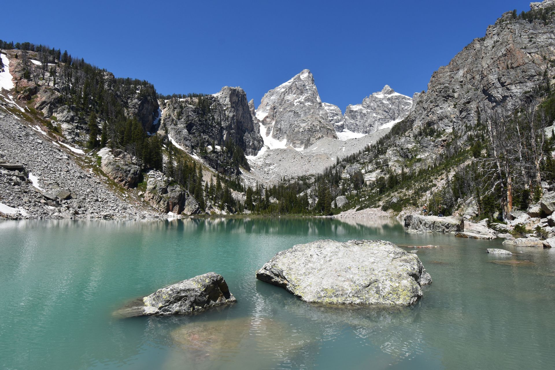 An aqua blue lake with pointed mountains in the distance. There are a couple large boulders in the lake.