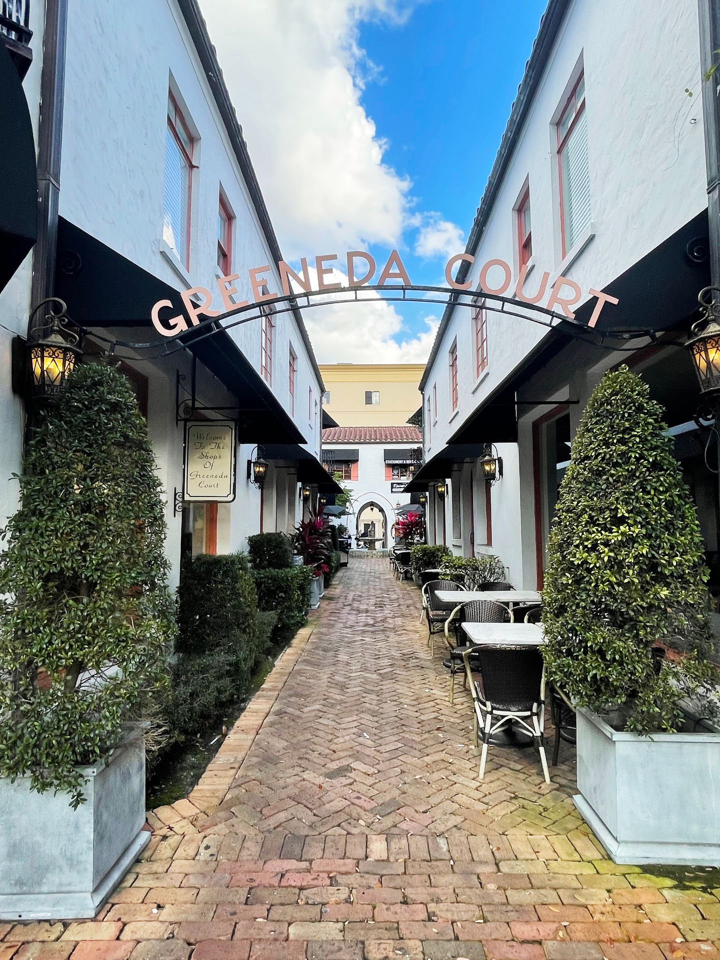 A alley way with white buildings on each side and the back. An arch above the alley reads "Greeneda Court." A few tables are on the side of the path.