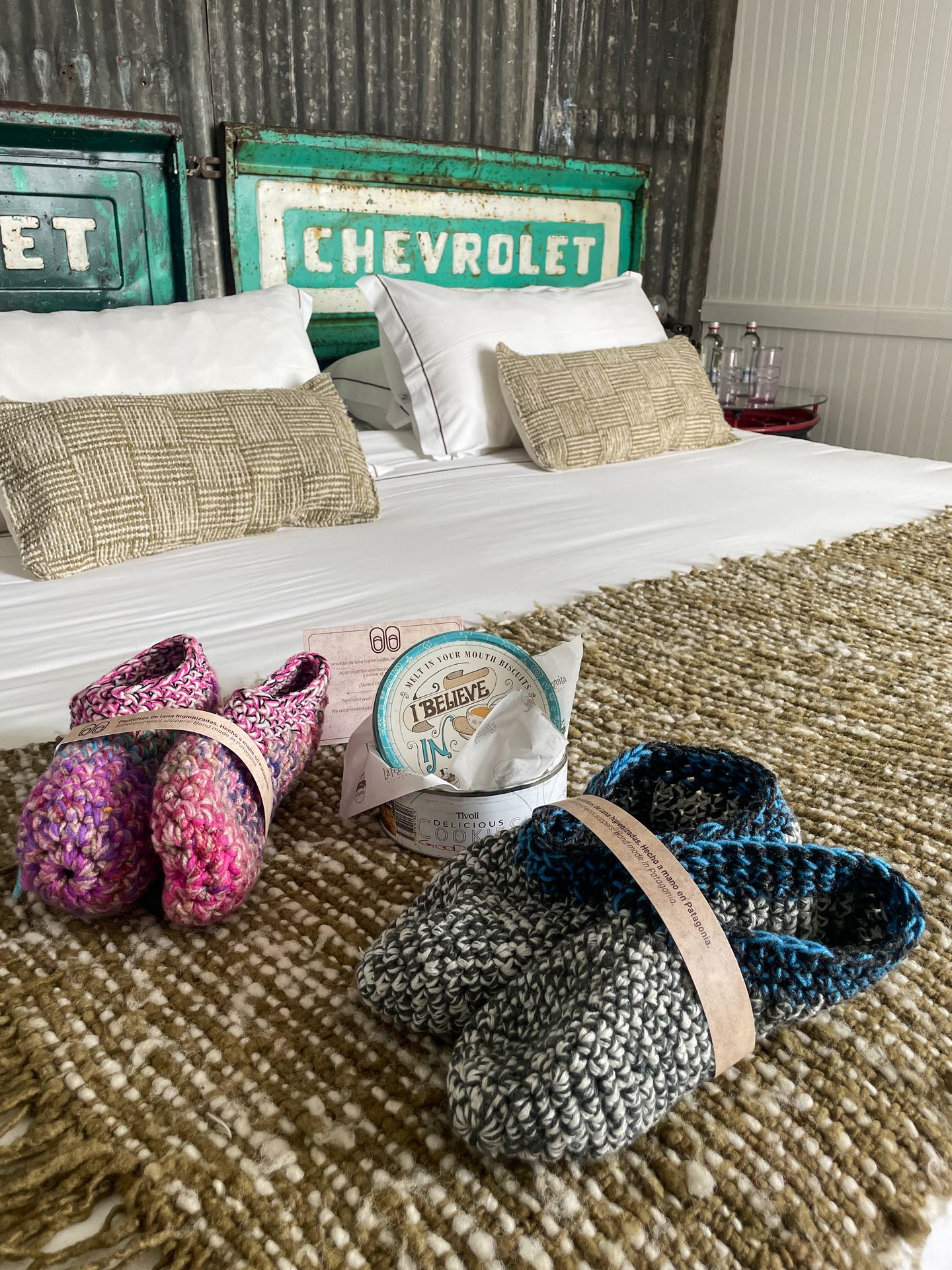 Two pairs of slipper and a tin of chocolate sitting on a bed. Behind the pillows is a piece of vintage sign that reads 'Chevrolet'