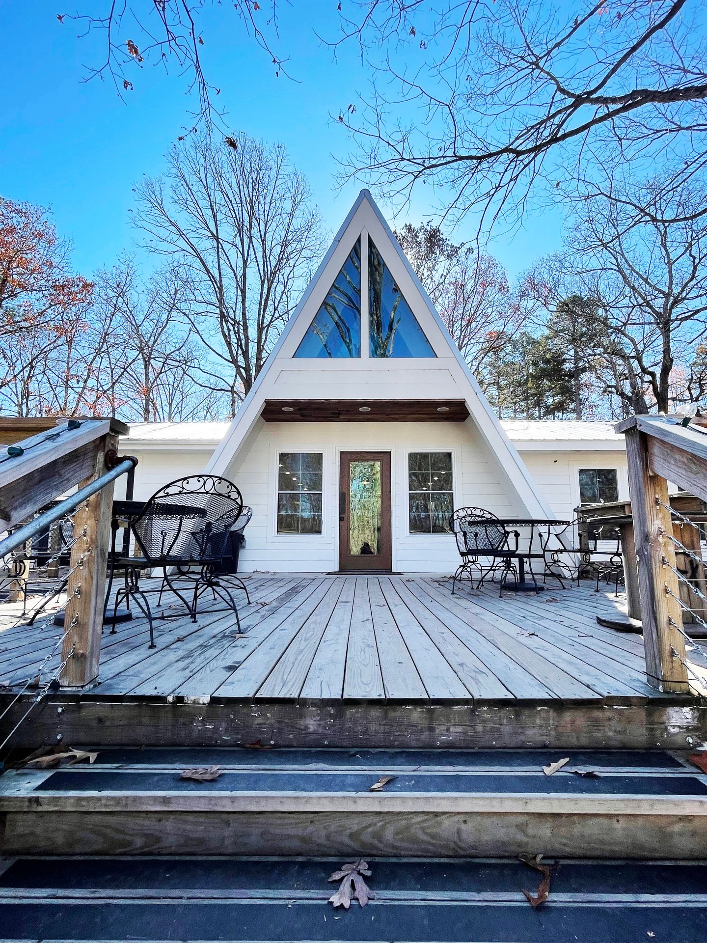 A white A-frame building with a deck in front. There are windows in the top of the A-frame and tables and chairs outside.