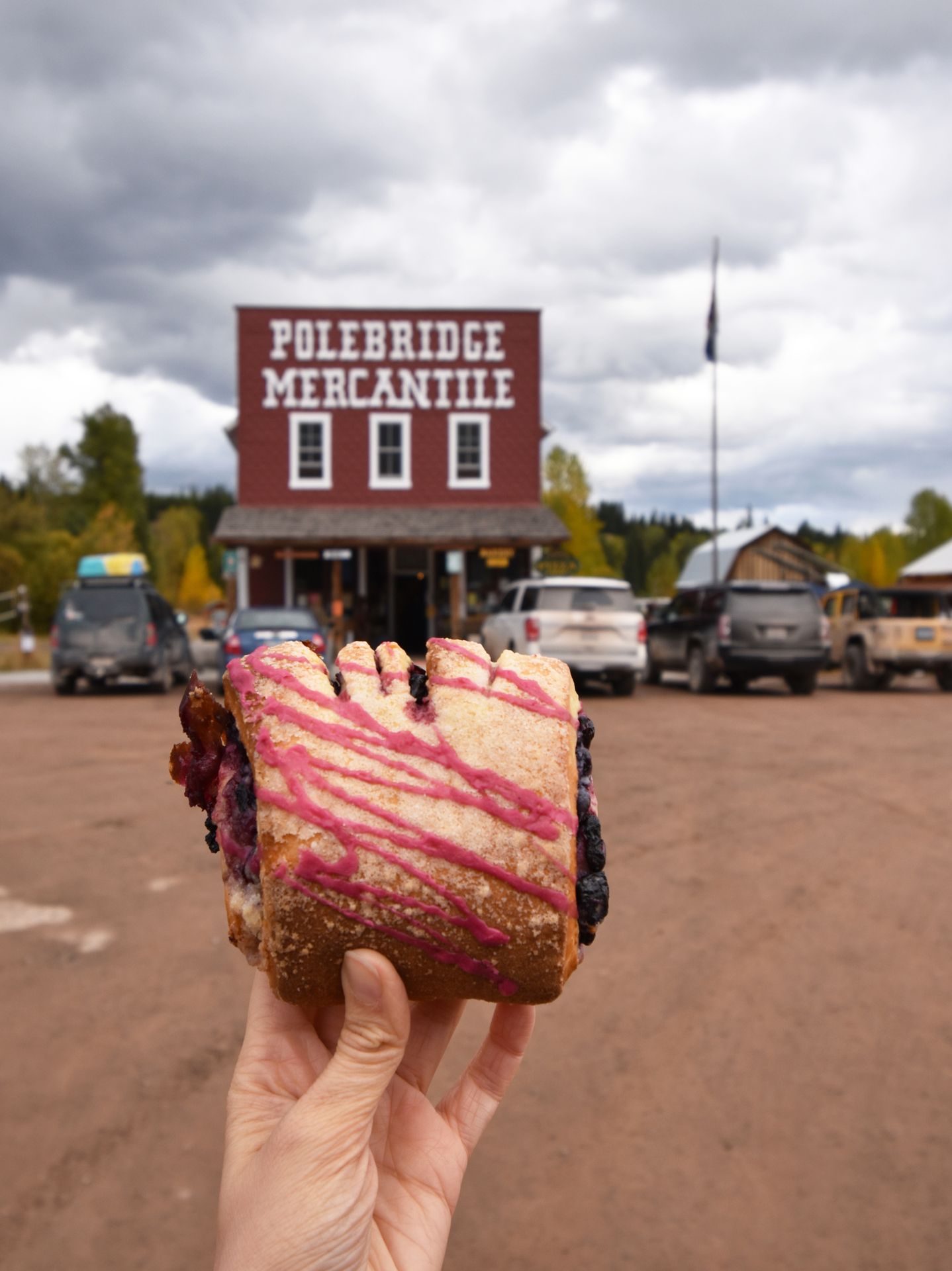 Holding up a huckleberry bearclaw in front of Polebridge Mercantile. The bearclaw has purple icing and Polebridge Mercantile is a red building with white letters.