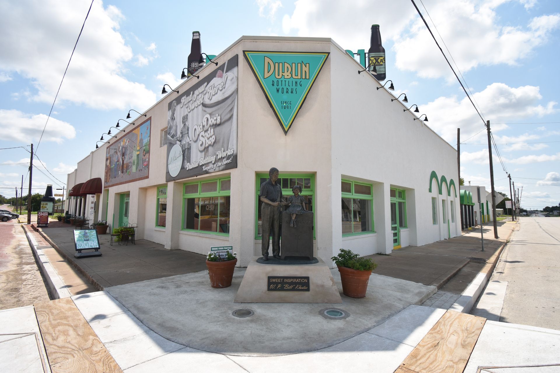 The exterior of Dublin Bottling Works front the corner. There is a statue in front of the building of W. P. Kloster, a triangle sign that reads "Dublin Bottling Works," a banner for Old Doc's Soda Shop and an empty sidewalk.