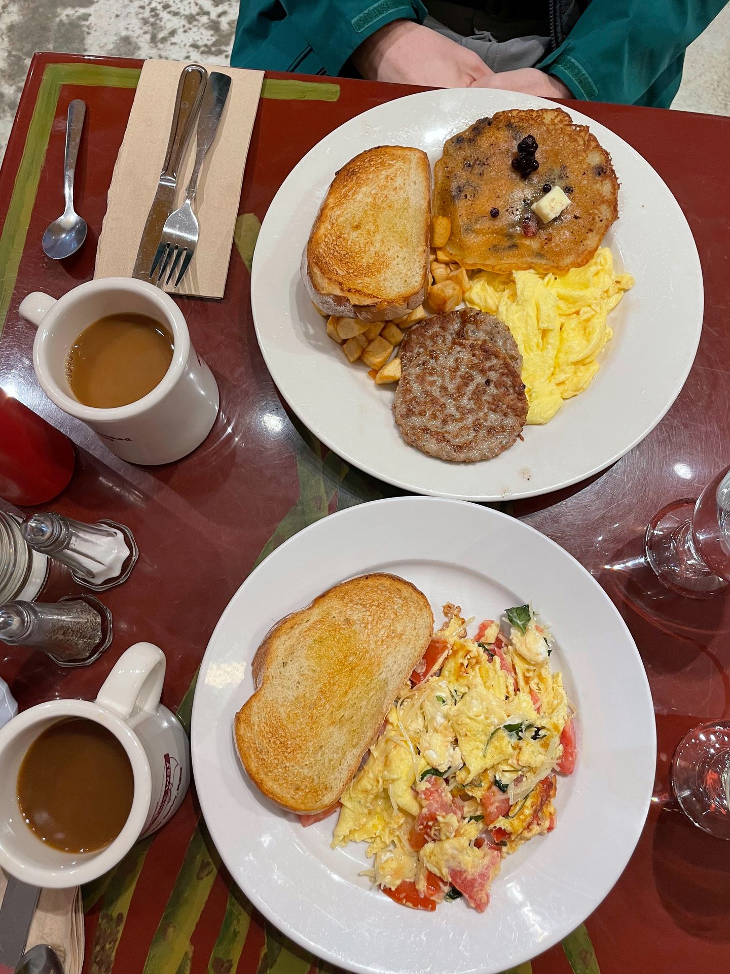 Two plates of breakfast food from Cafe This Way. One has am scrambled eggs with veggies and toast. The other has pancakes, scrambled eggs, sausage and toast.