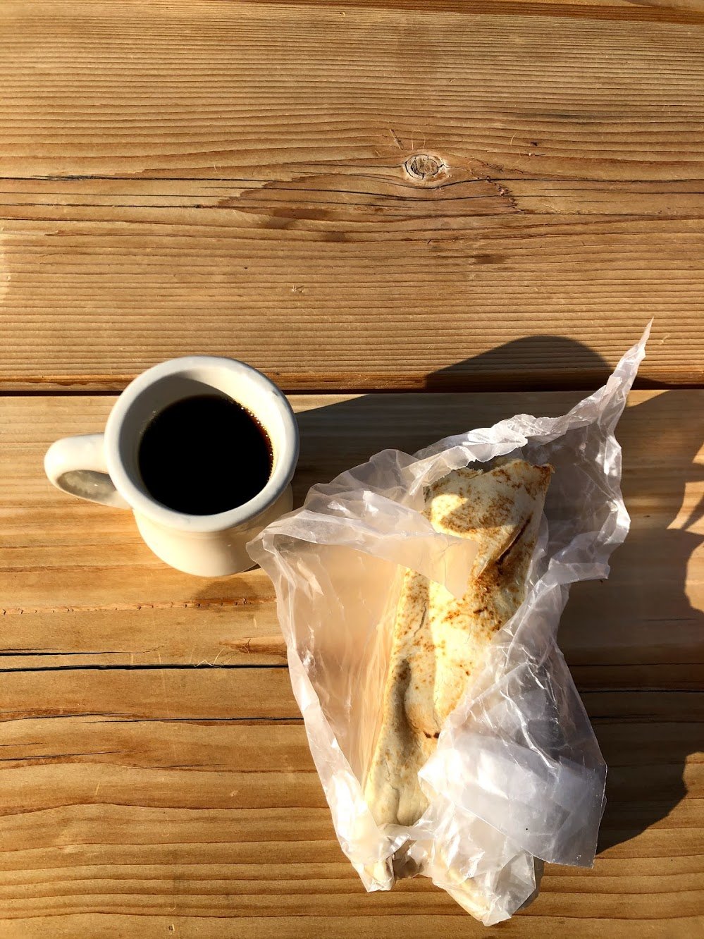 A large burrito sitting on a table, loosely wrapped in paper. A mug of coffee sits next to it.