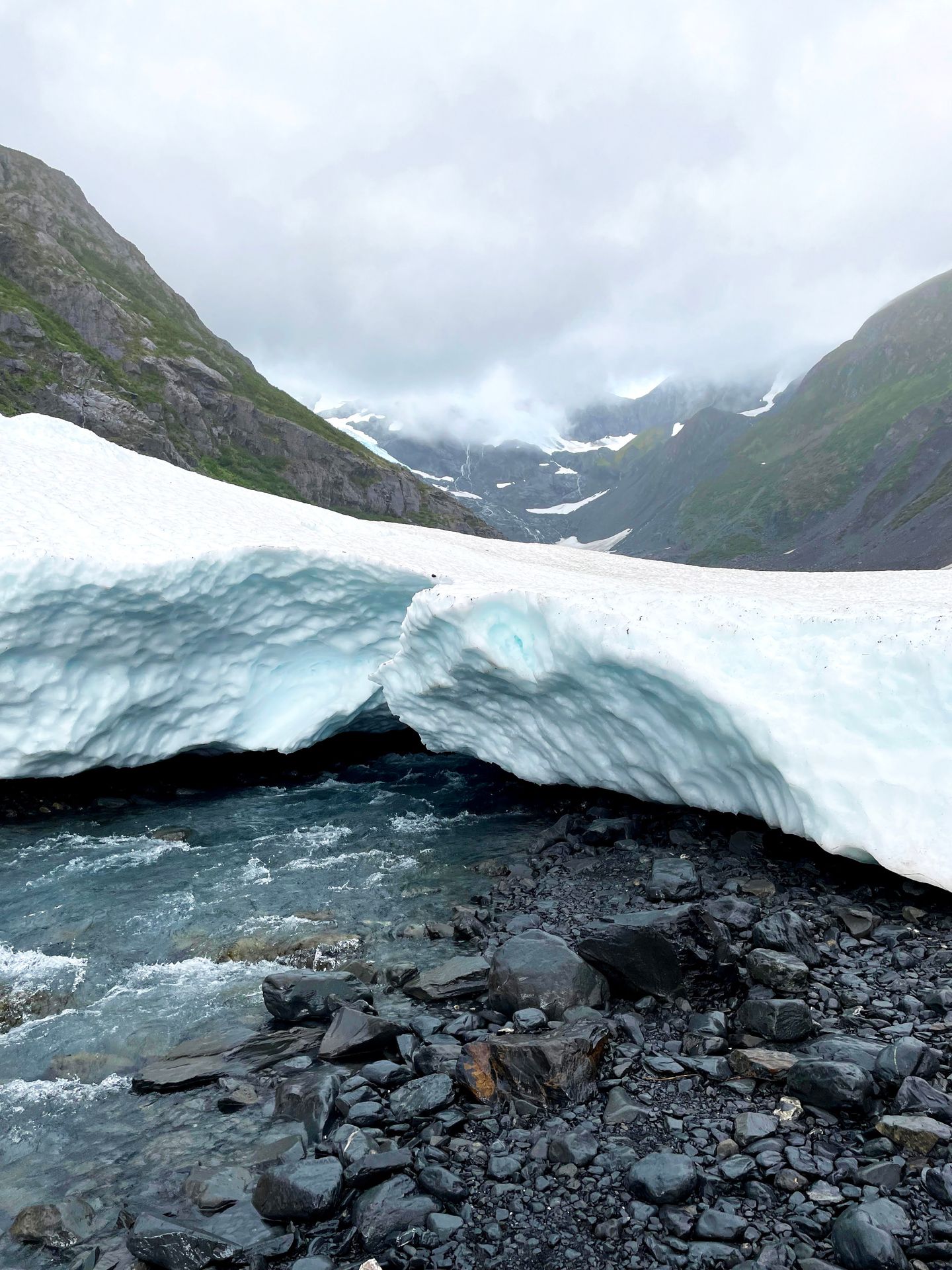A small glacier with a river flowing under it and mountains in the background.