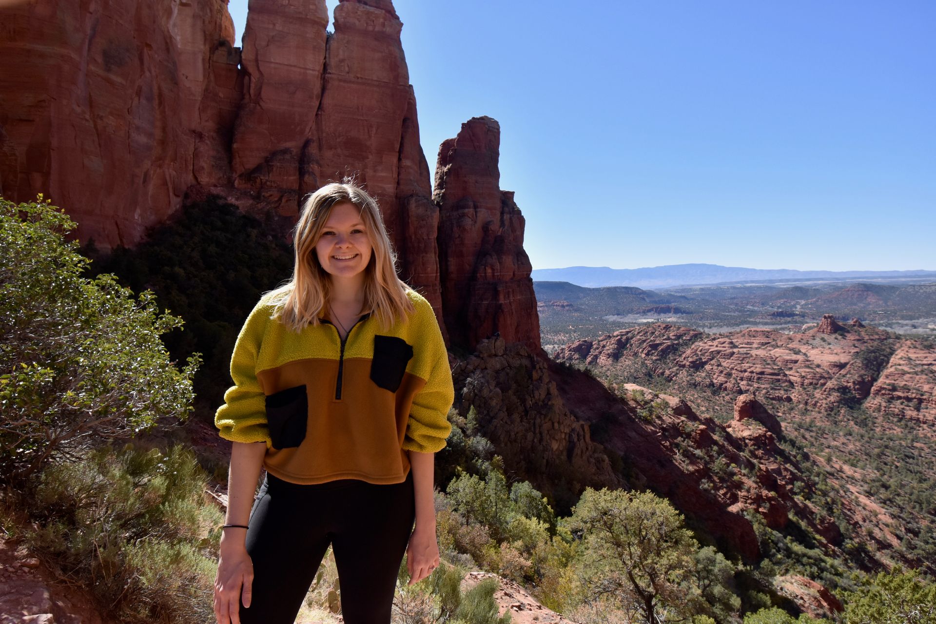 Lydia standing at the viewpoint on the Cathedral Rock trail. She is wearing a yellow and orange color block sweathshirt.