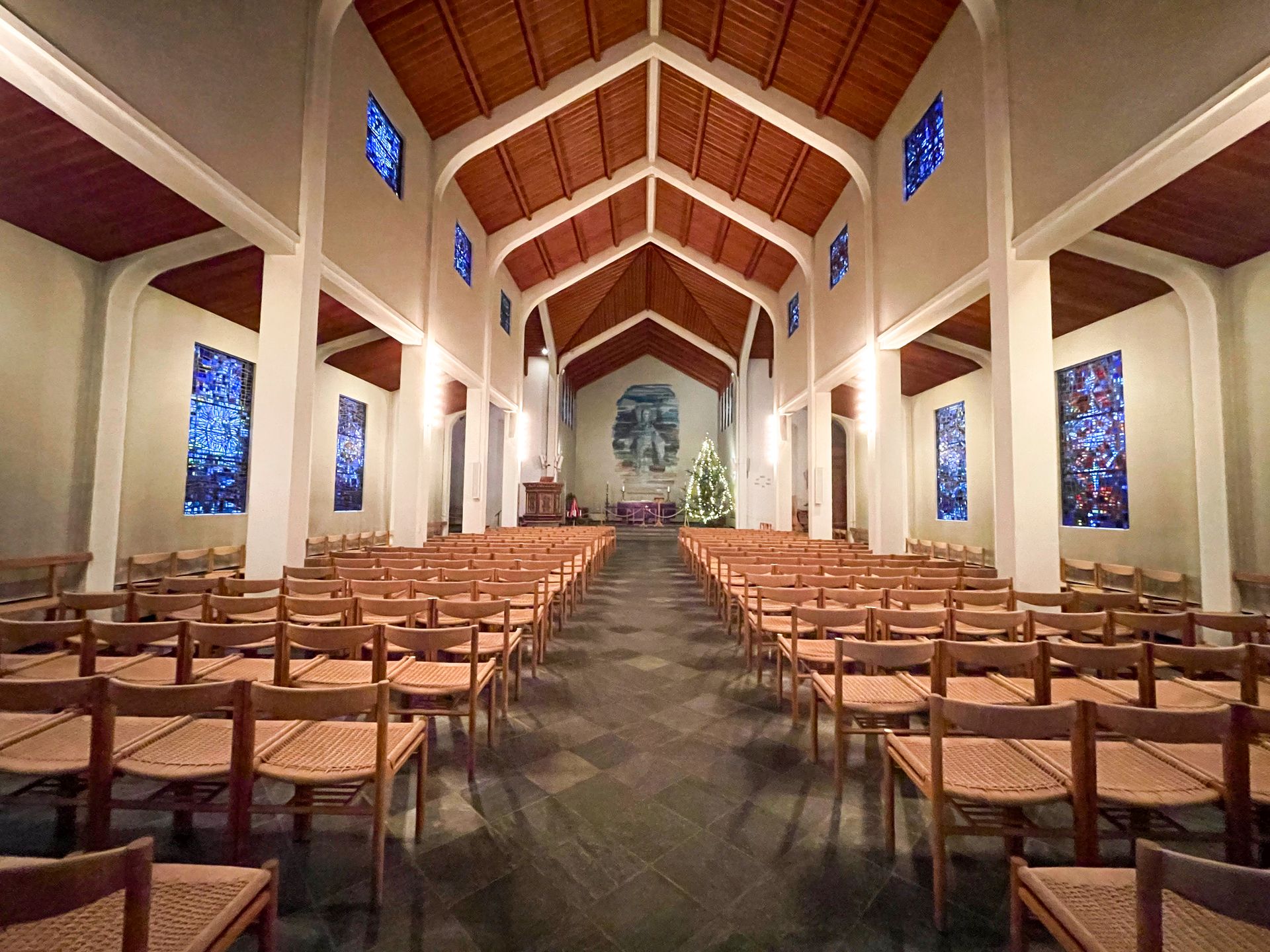 The interior of Skalholt Church.  The walls are white with a brown ceiling and brown chairs. There are stained glass windows on the walls.