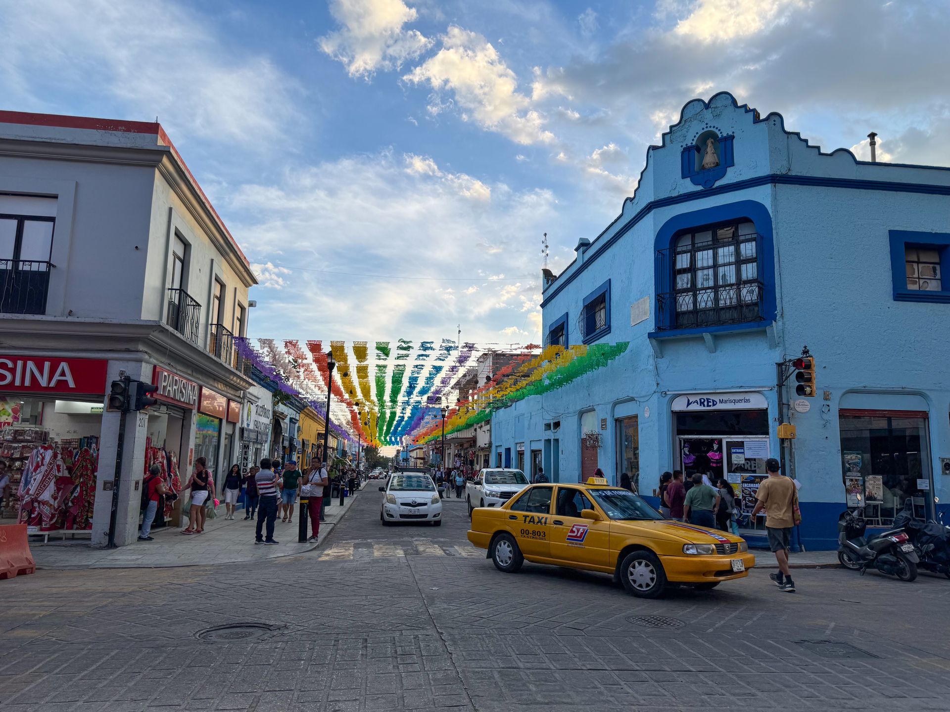 A blue building, a yellow taxi and a street that has rainbow flags hanging over it