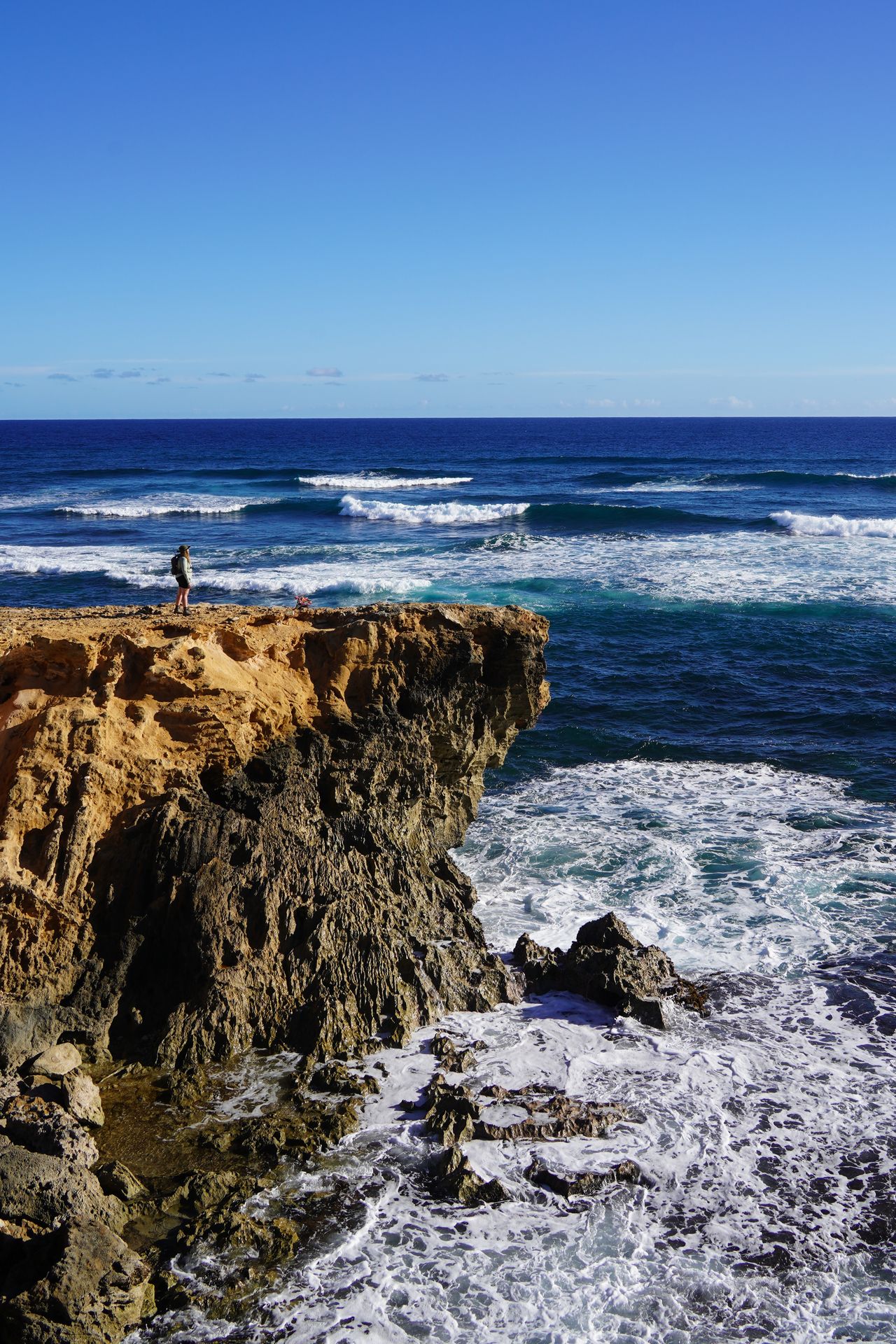 Lydia standing far away on a yellow cliff that stands up above a ocean with several waves