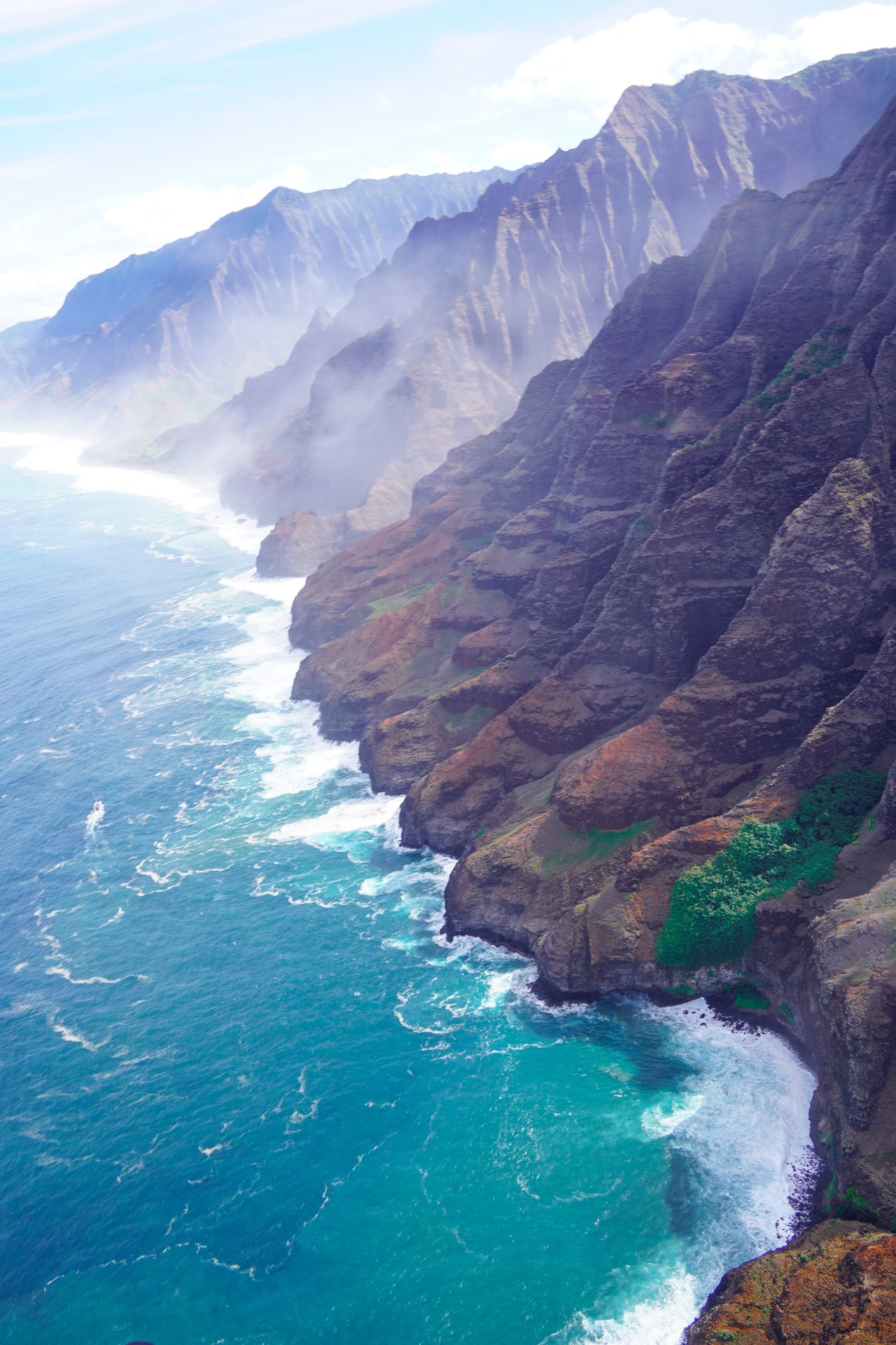 A view of the Na Pali Coast from above. The ocean is an aqua blue and there is some mist hanging next to the cliffs