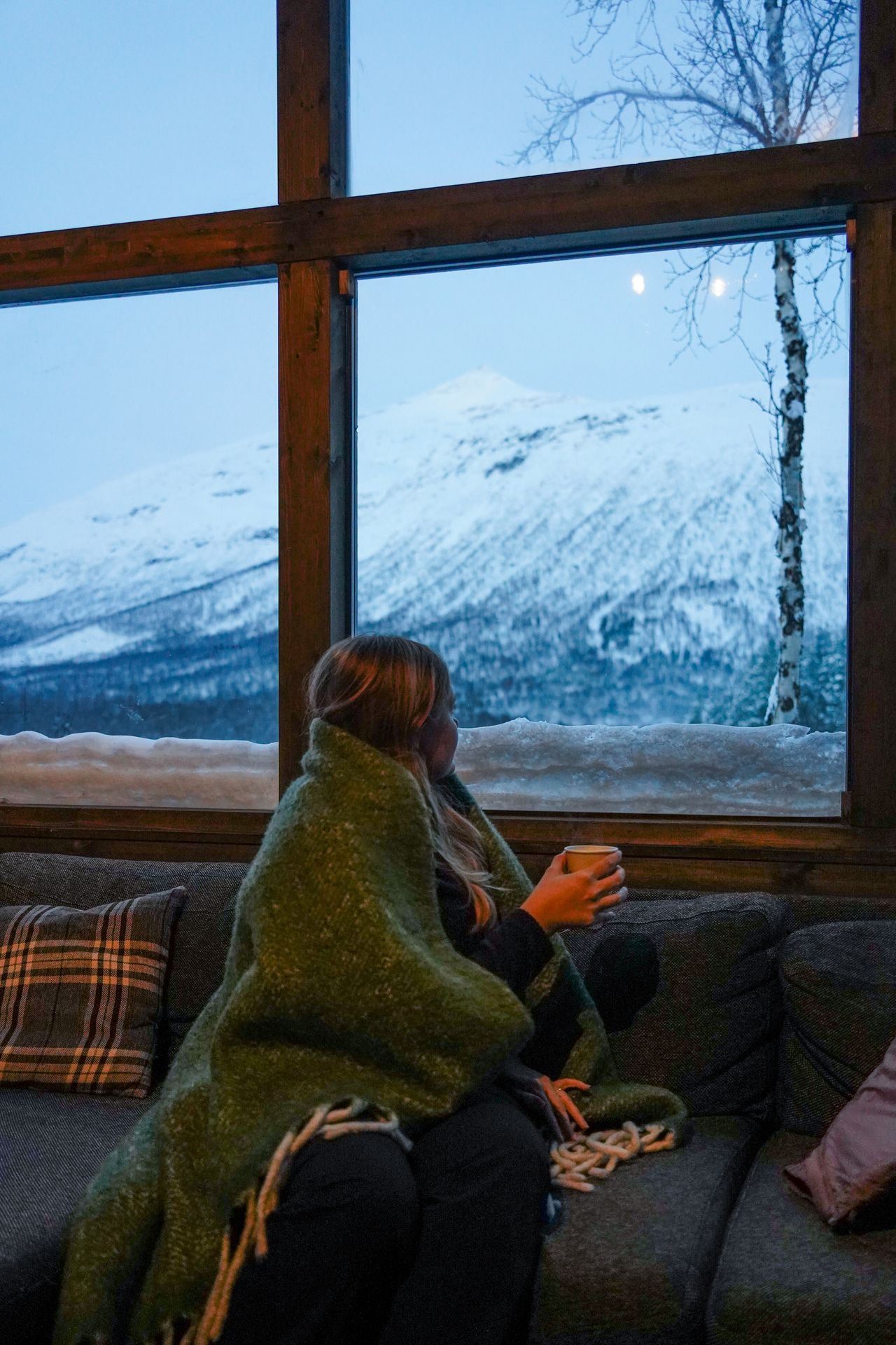 Lydia looking out a large window at snow. She is under a green blanket and holds a coffee