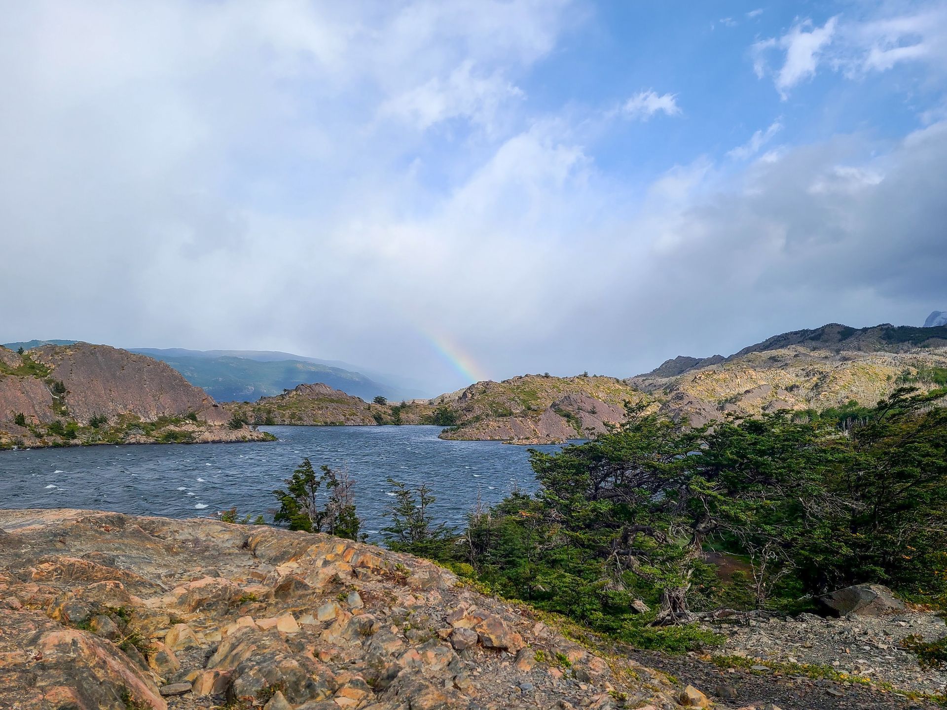 A blue lake surrounded by mountains and hills of various sizes. There is a small rainbow coming up from one of the hills.