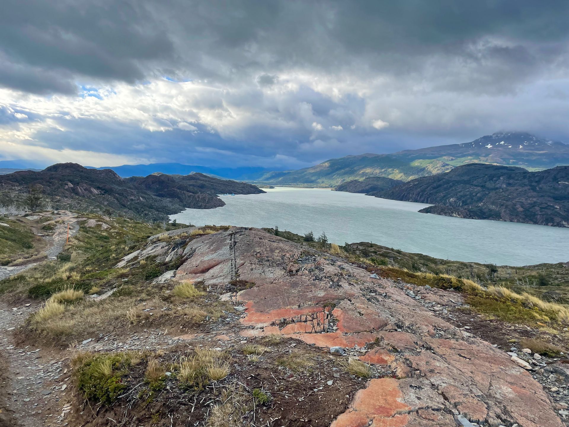 A large glacier-fed lake is in the distance. In the foreground, there are some reddish rocks. There are green mountains in the distance behind the lake.