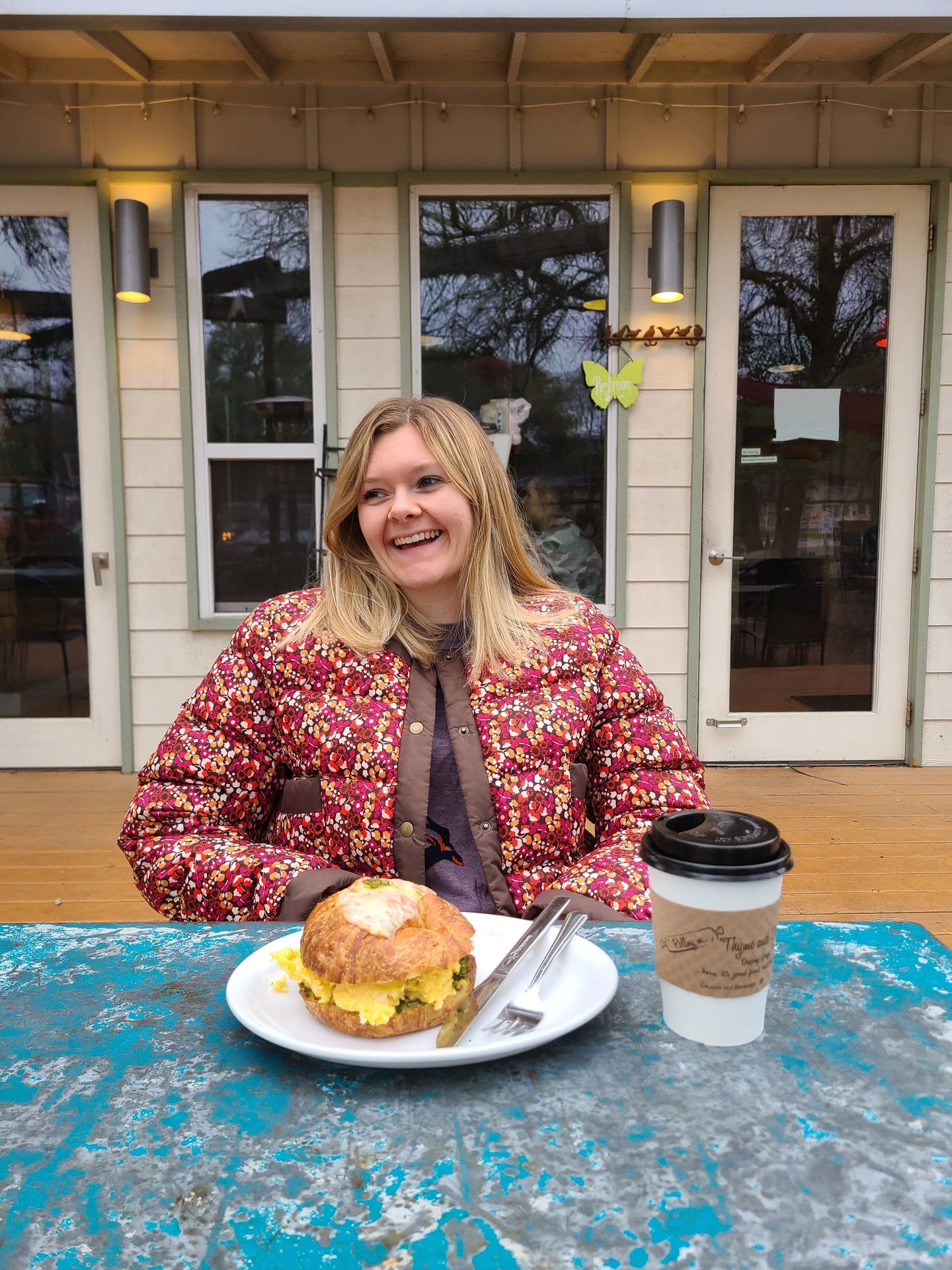 Lydia sitting at a table at Rolling in Thyme and Dough. A giant croissant sandwich with scrambled eggs sits on the table.