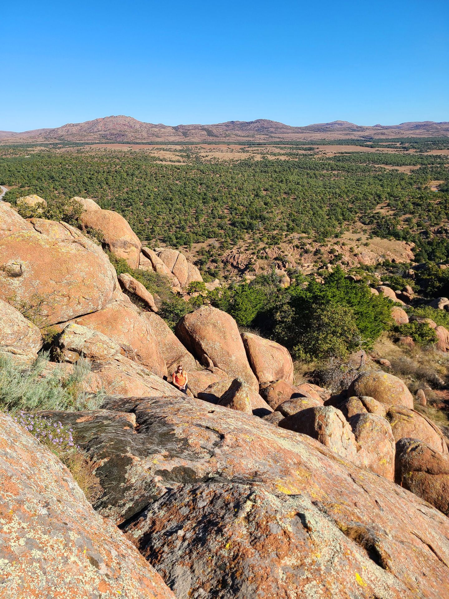 Large orange boulders with a view of greenery in the distance. Lydia sits on the rocks but is hard to see.