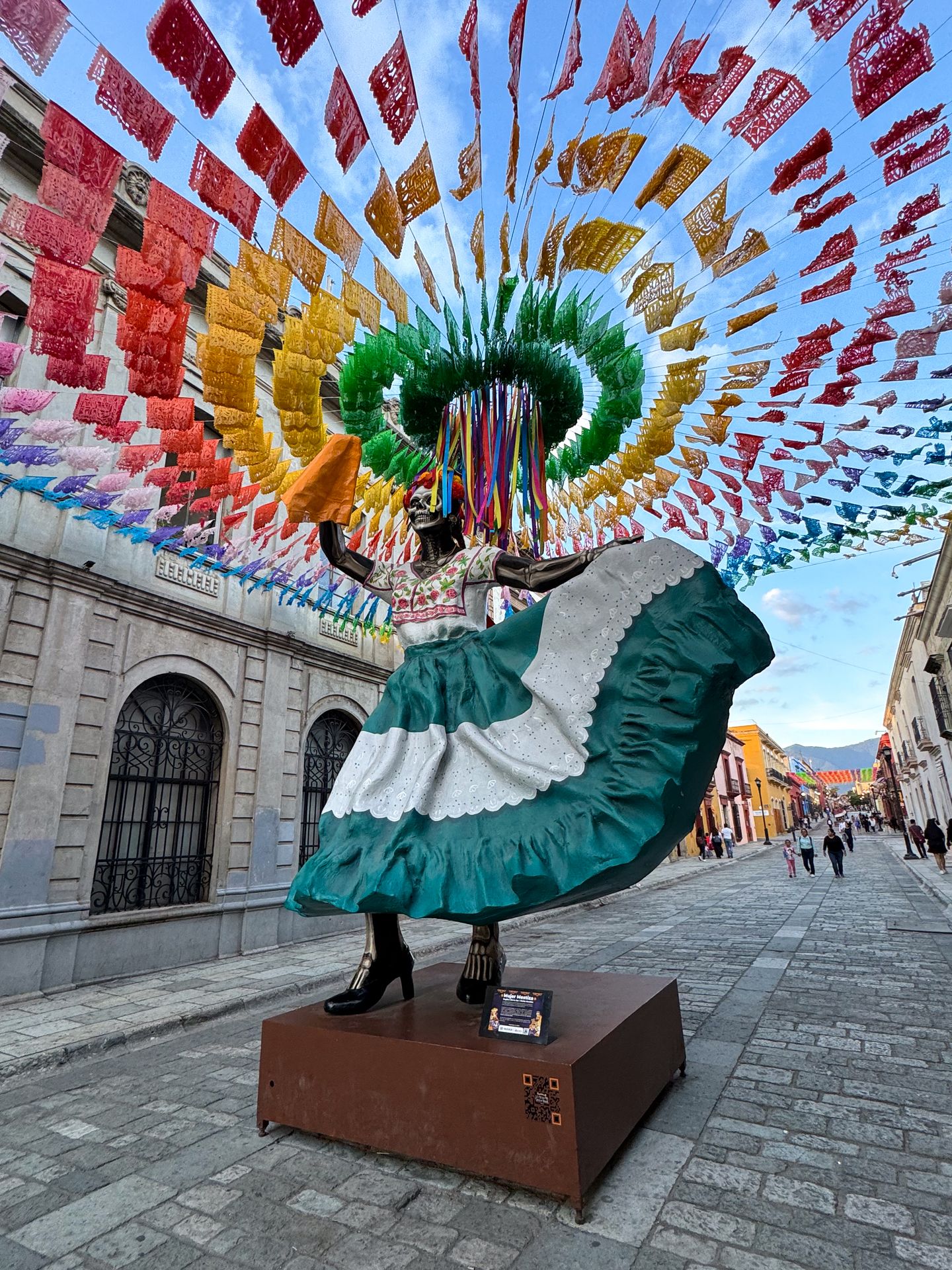 A statue of a woman in a skirt in the middle of the street, with rainbow flags above