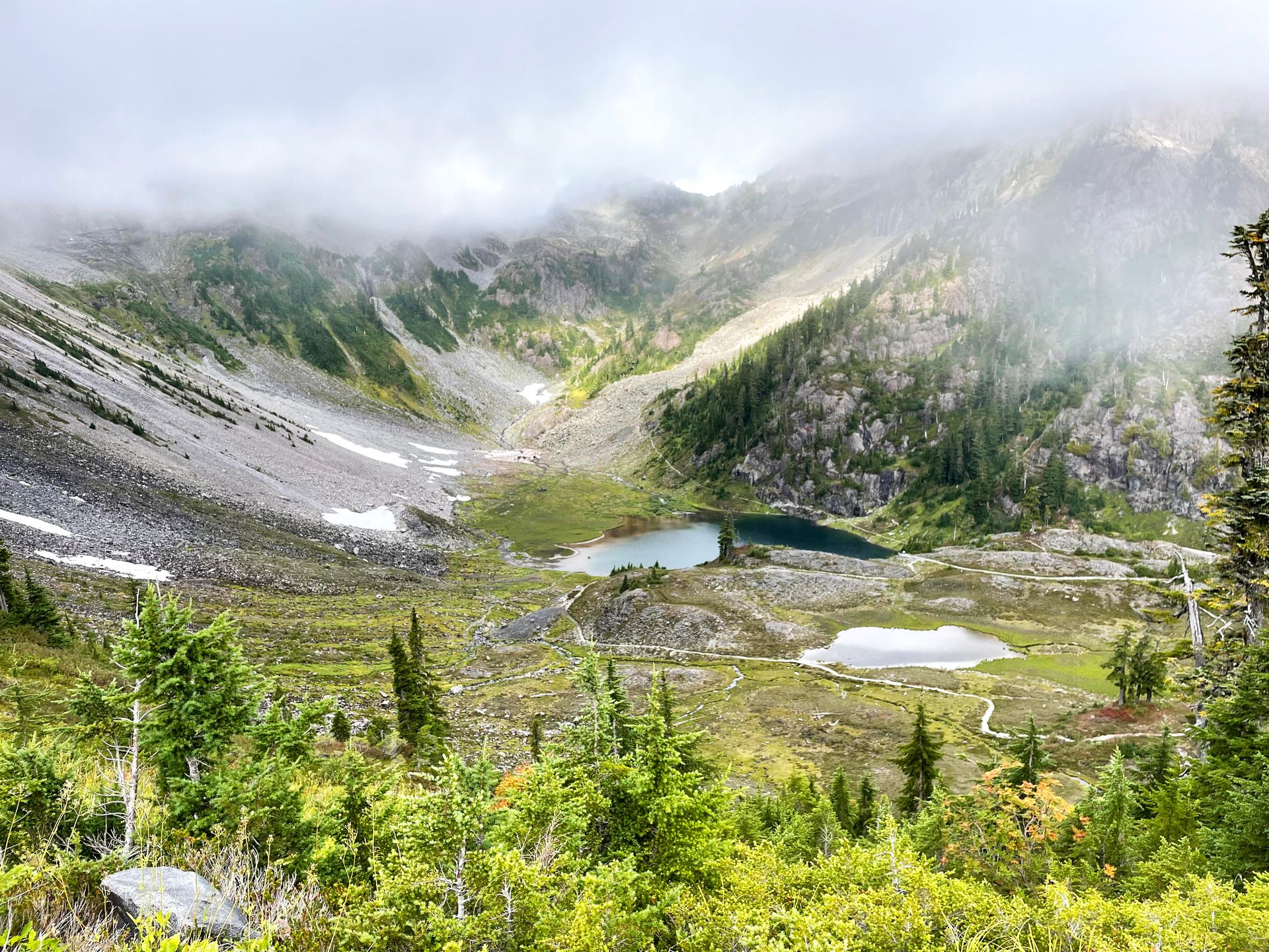 An area of mountains, green trees and a small lake with fog in the air.