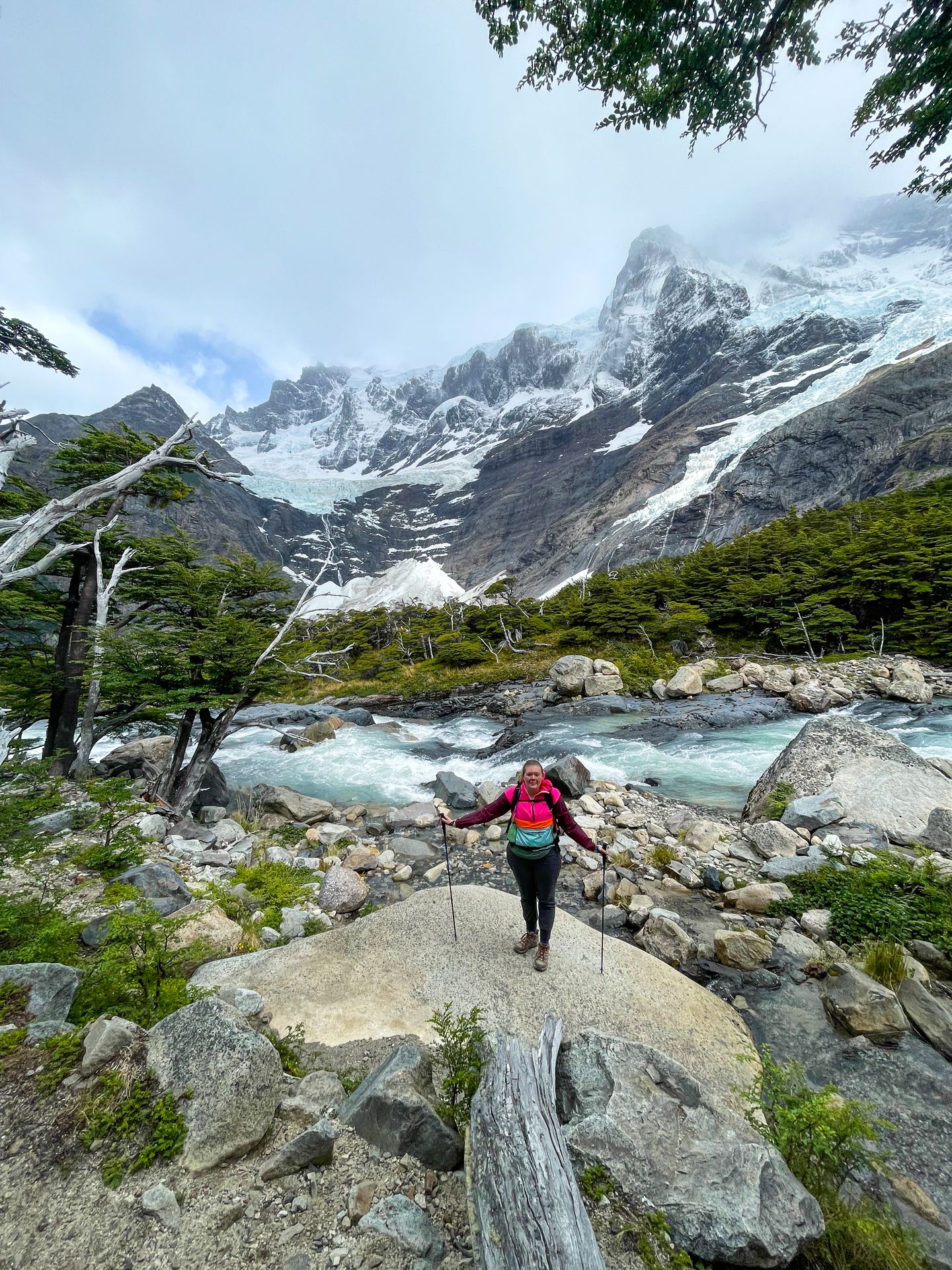 Lydia standing on a large rock and holding out her trekking poles. In the background is a blue river with mountains in the distance.