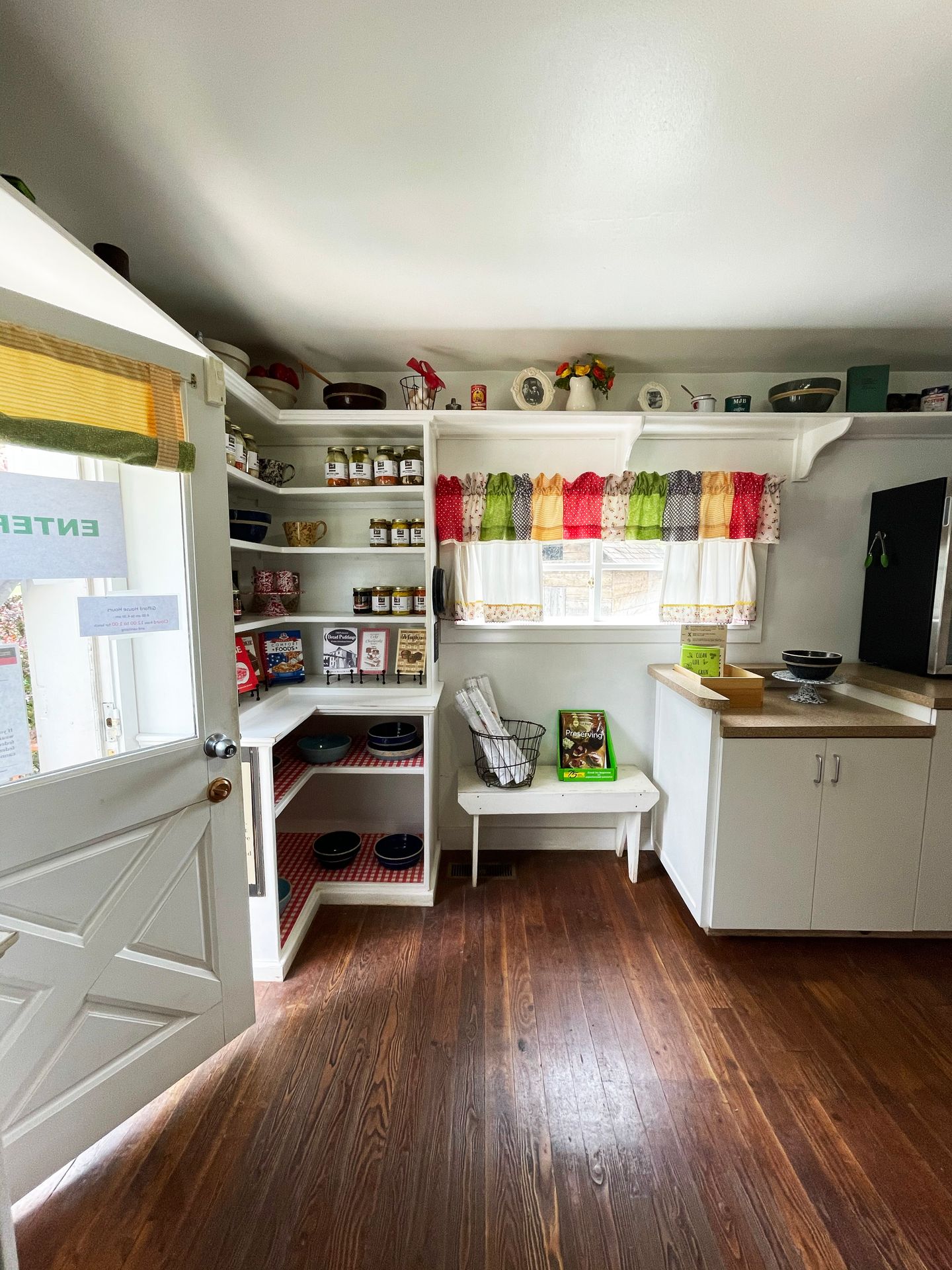 The inside of a shop with white walls and a wood floor. There are colorful curtains and jars on the walls.