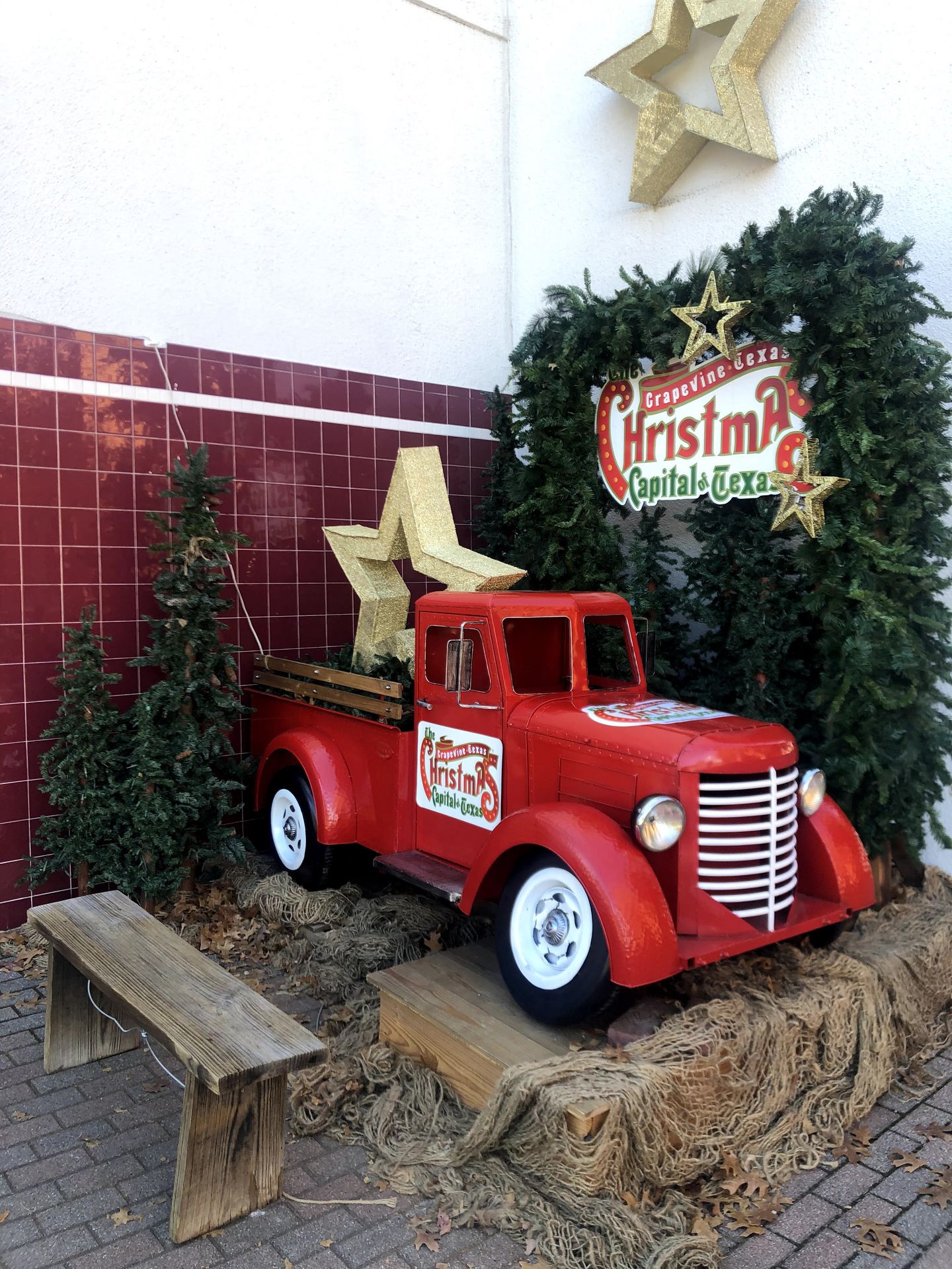 A mini red pick up truck with a gold star in the truck bed. It sits on a bail of hay in front of a background made of green pines.