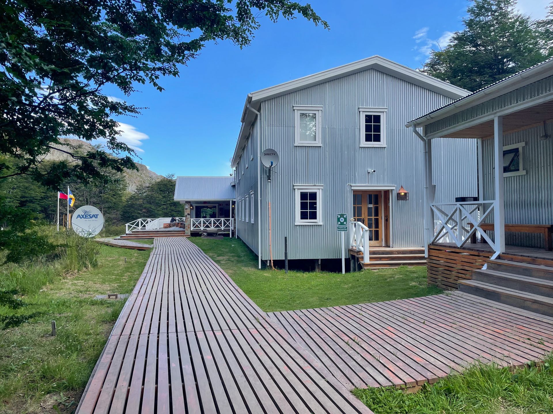 A couple of light blue buildings with a boardwalk trail connecting them. There is a porch on each buildings and a satellite dish outside.