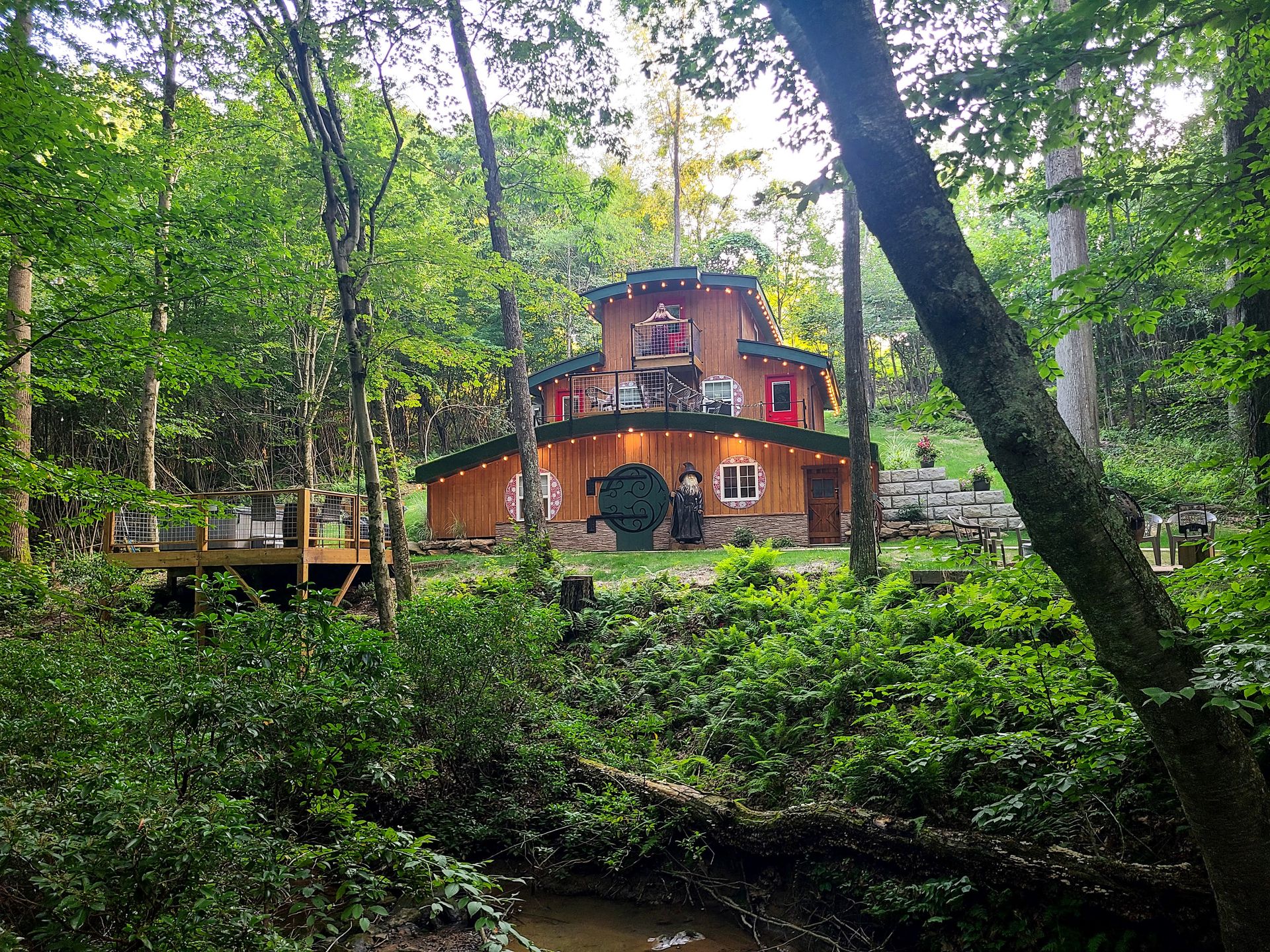 Lydia standing on the 3rd floor balcony at the Ohiopyle Hobbit House. The home is brown with a green roof and 3 red doors. In front of the house is a hot tub and a creek.