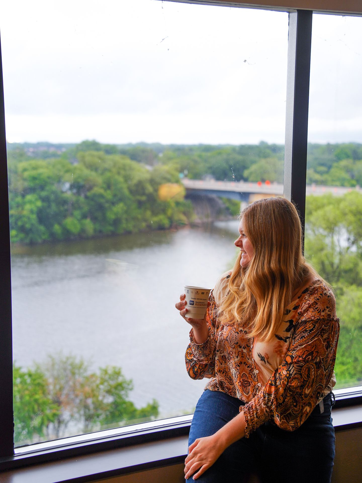 Lydia sitting by a window holding a cup of coffee in the Best Western Kelly Inn
