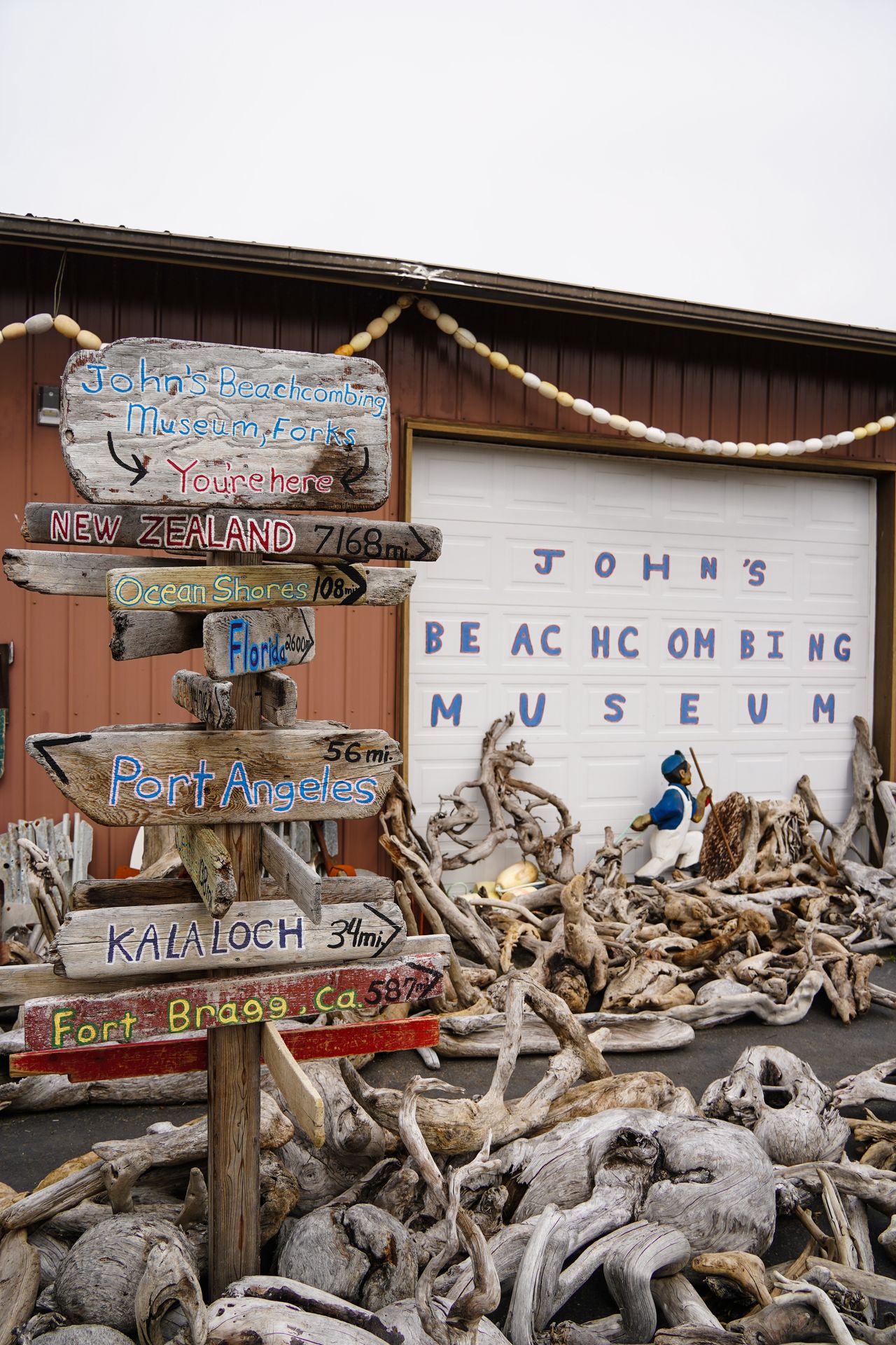A collection of driftwood, a garage door that reads 'John's Beachcombing Museum' and a sign that has the distances to several nearby towns, such as Port Angeles