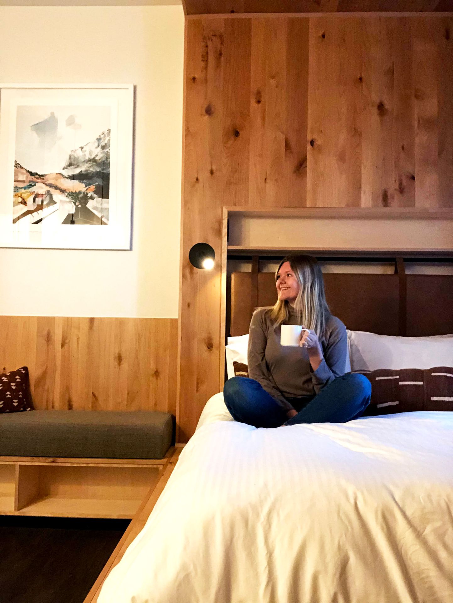 Lydia sitting on a bed and holding a coffee inside of Kinship Landing. There wood on the wall and white bedding.