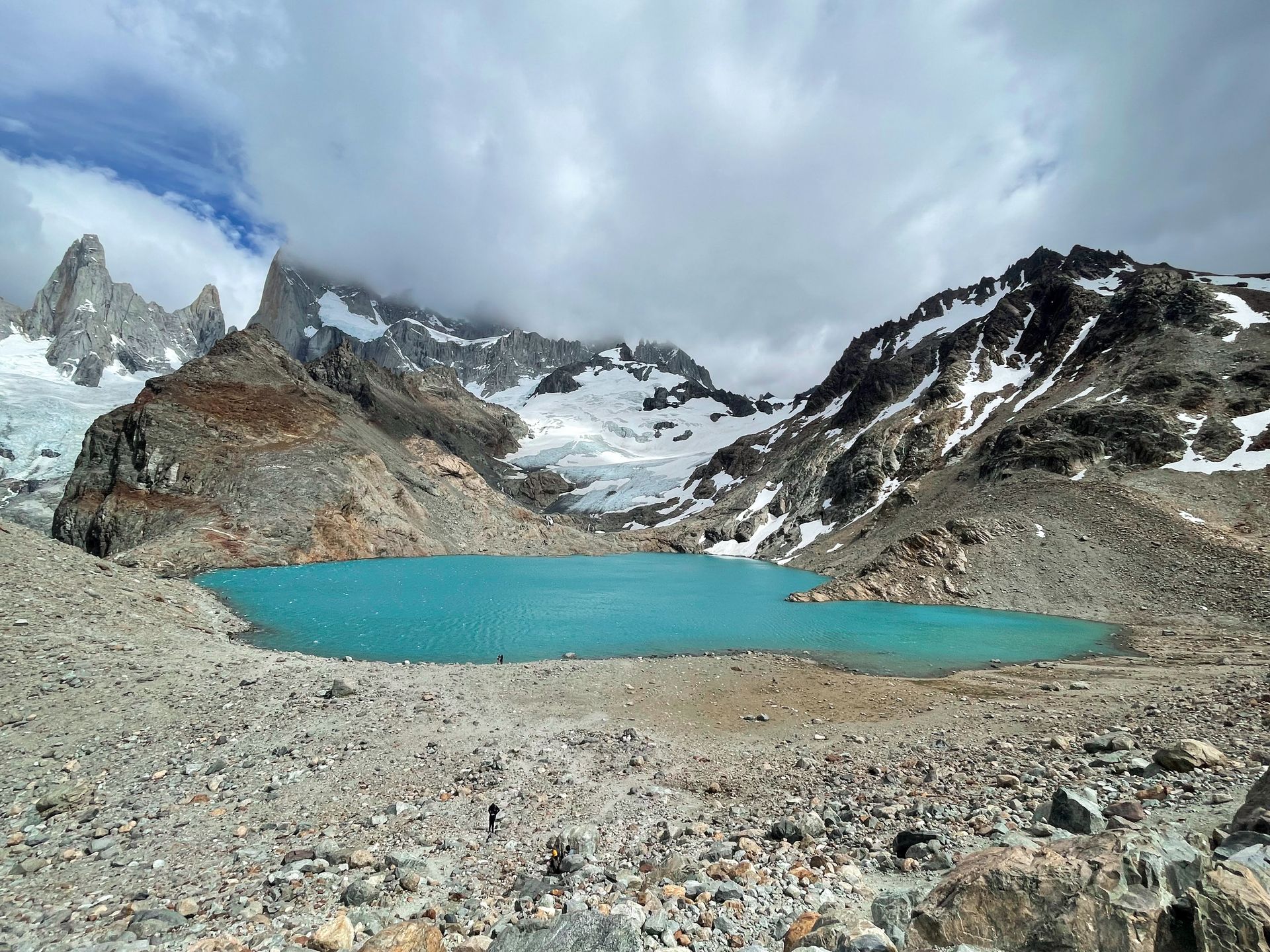 A small, bright blue lake with mountains in the background. The top of the mountains are obscured by clouds. There are multiple glaciers hanging on the mountains.
