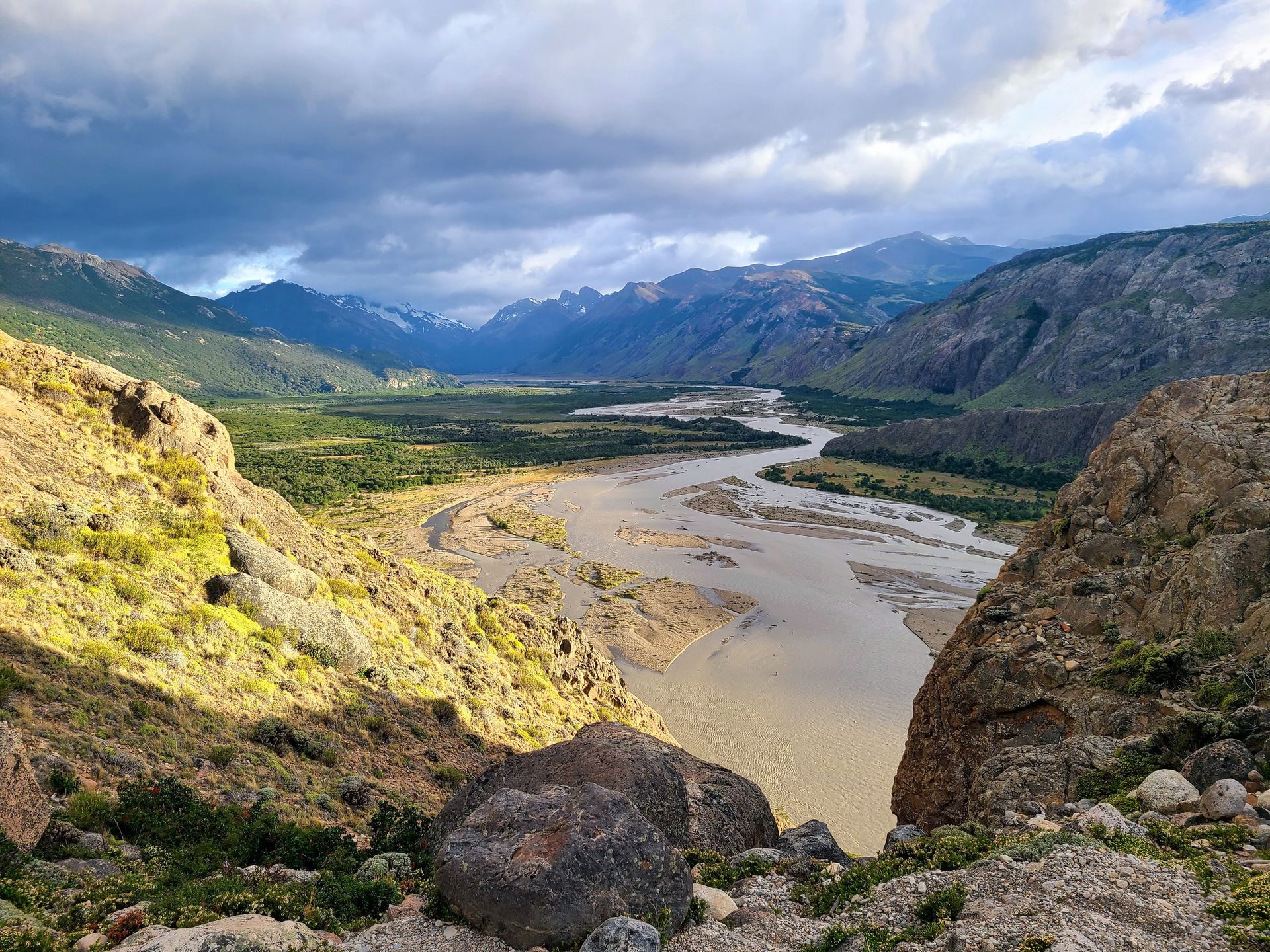Looking down at a wide river next to a green valley. The river is framed by mountains in the distance.