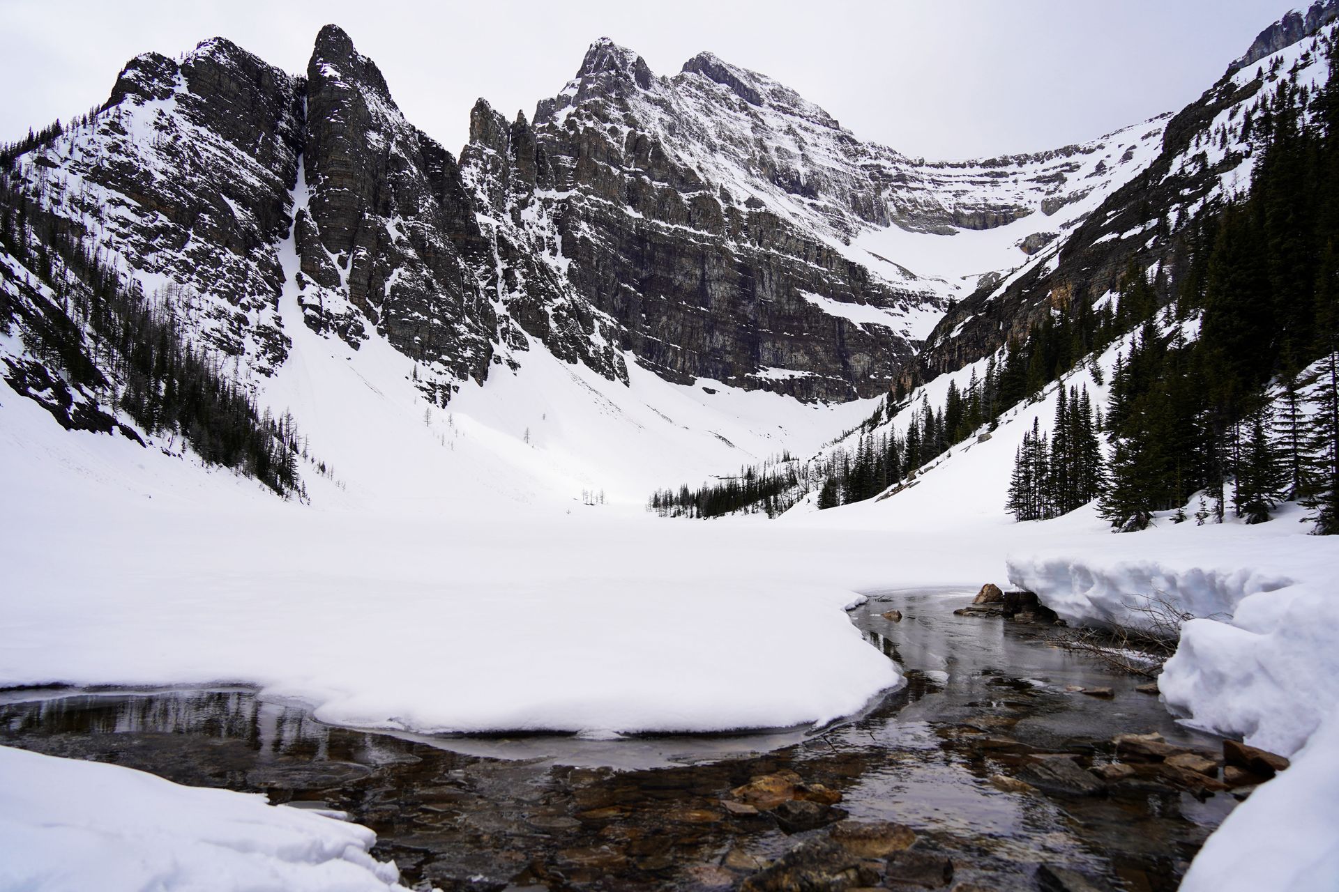 A partially frozen lake and mountains in the background. There is a couple feet of snow on top of the lake and mountains.