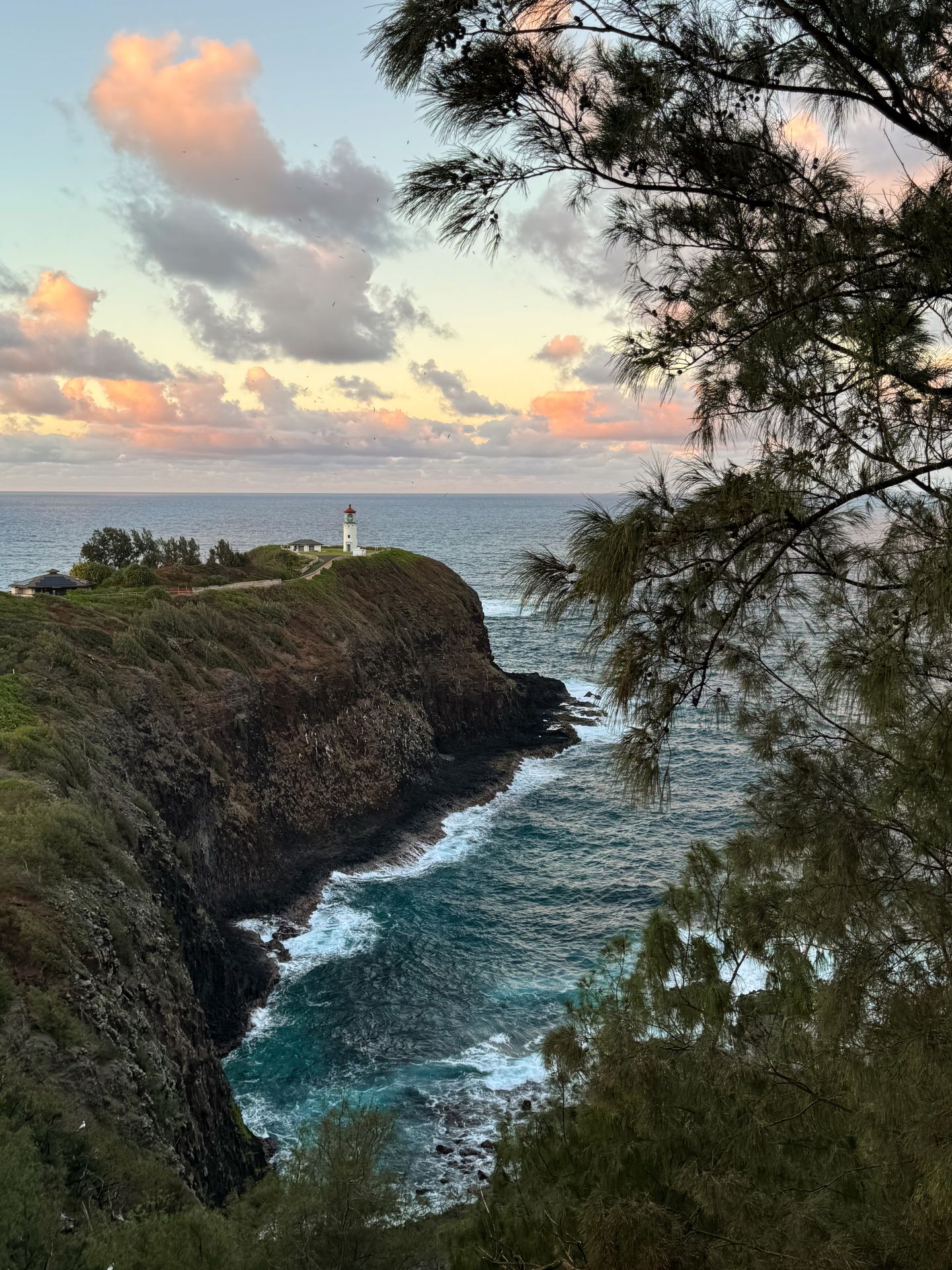 A lighthouse on the edge of a cliff that extends out into the ocean, with a pink sky at sunset