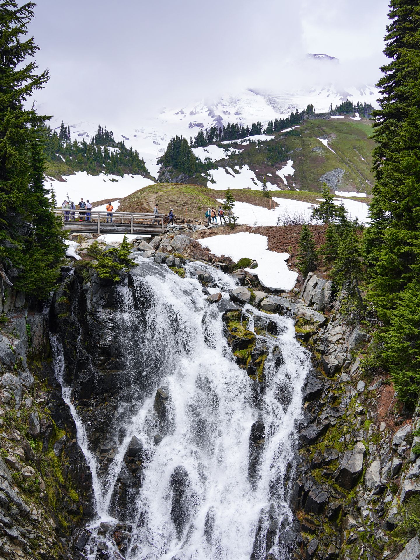 A waterfall that has a bridge over it, with people on the bridge. You can see Mount Rainier in the background, partially obscured by fog.
