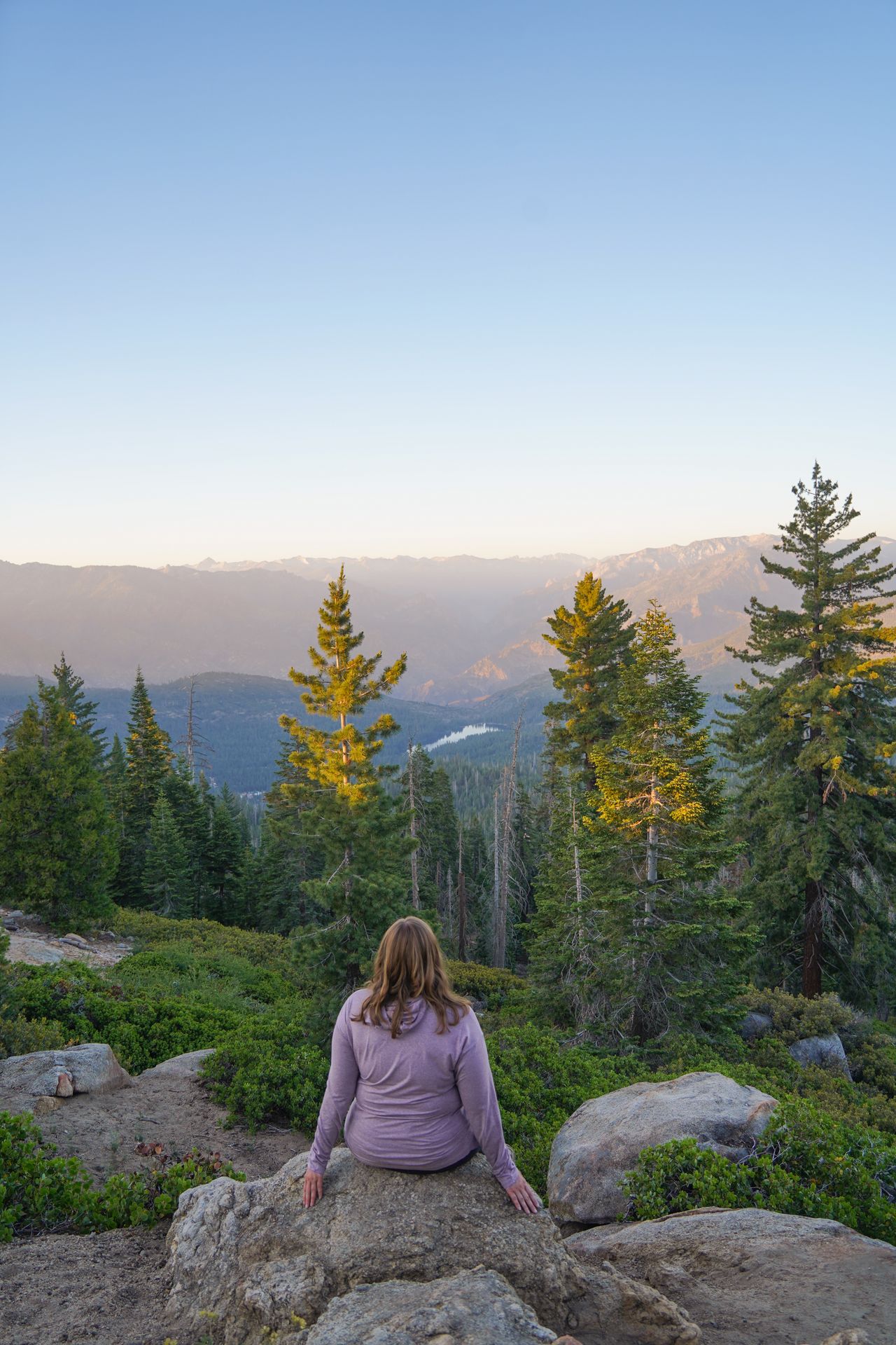 Lydia sitting on a rock and looking out at mountains, trees and a lake that are glowing pink at sunset