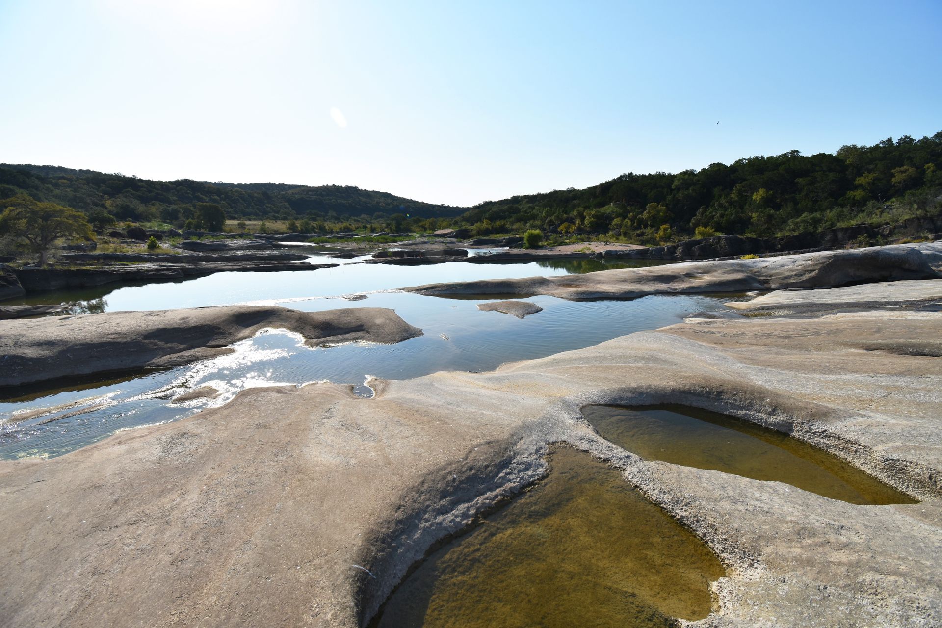 A view of Pedernales Falls with multiple pools of water in the various rock faces. In the distance, there are green trees.