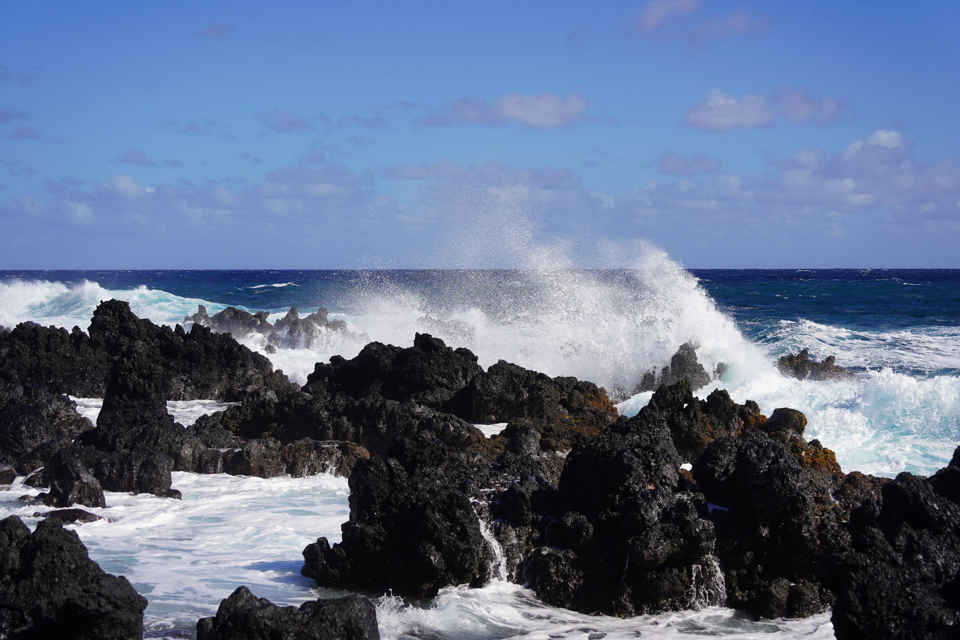 Waves crashing into a group of jagged black lava rocks on the coast