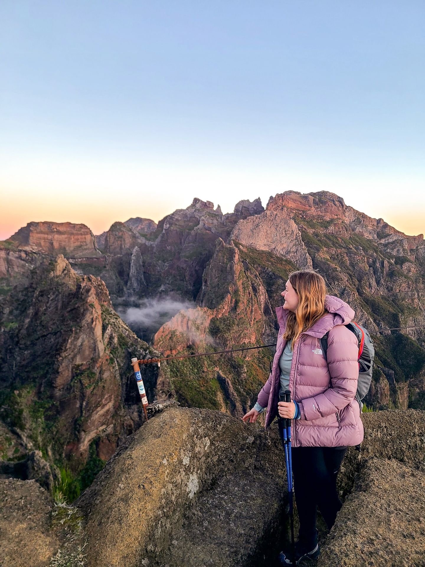 Lydia looking out at the view of mountains on the PR1 Trail. She wears a light purple coat and holds trekking poles.
