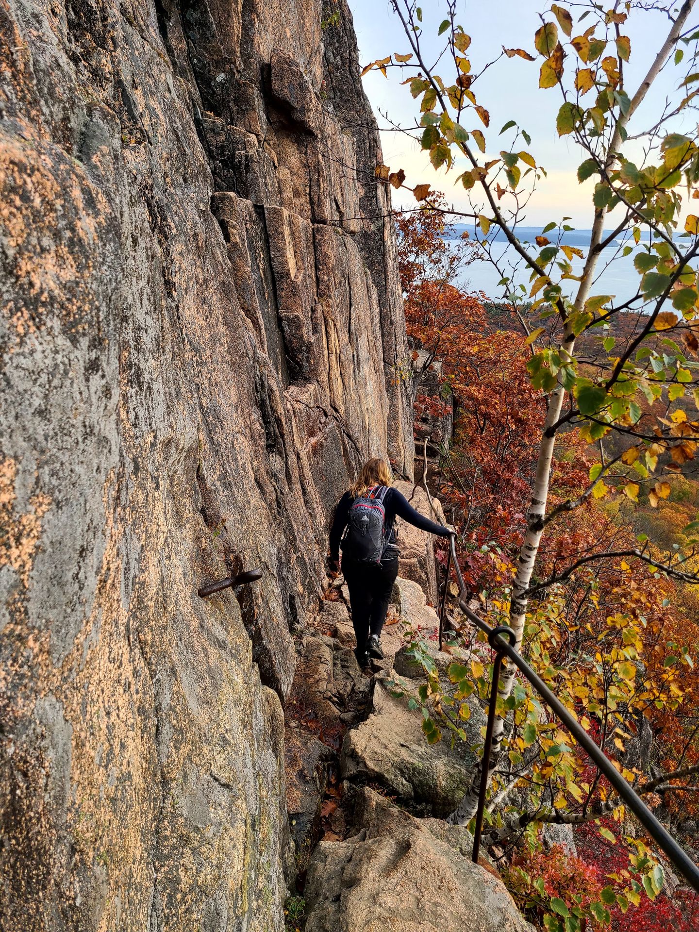 Lydia hiking on a narrow cliff path and touching a metal railing on the Precipice Trail.