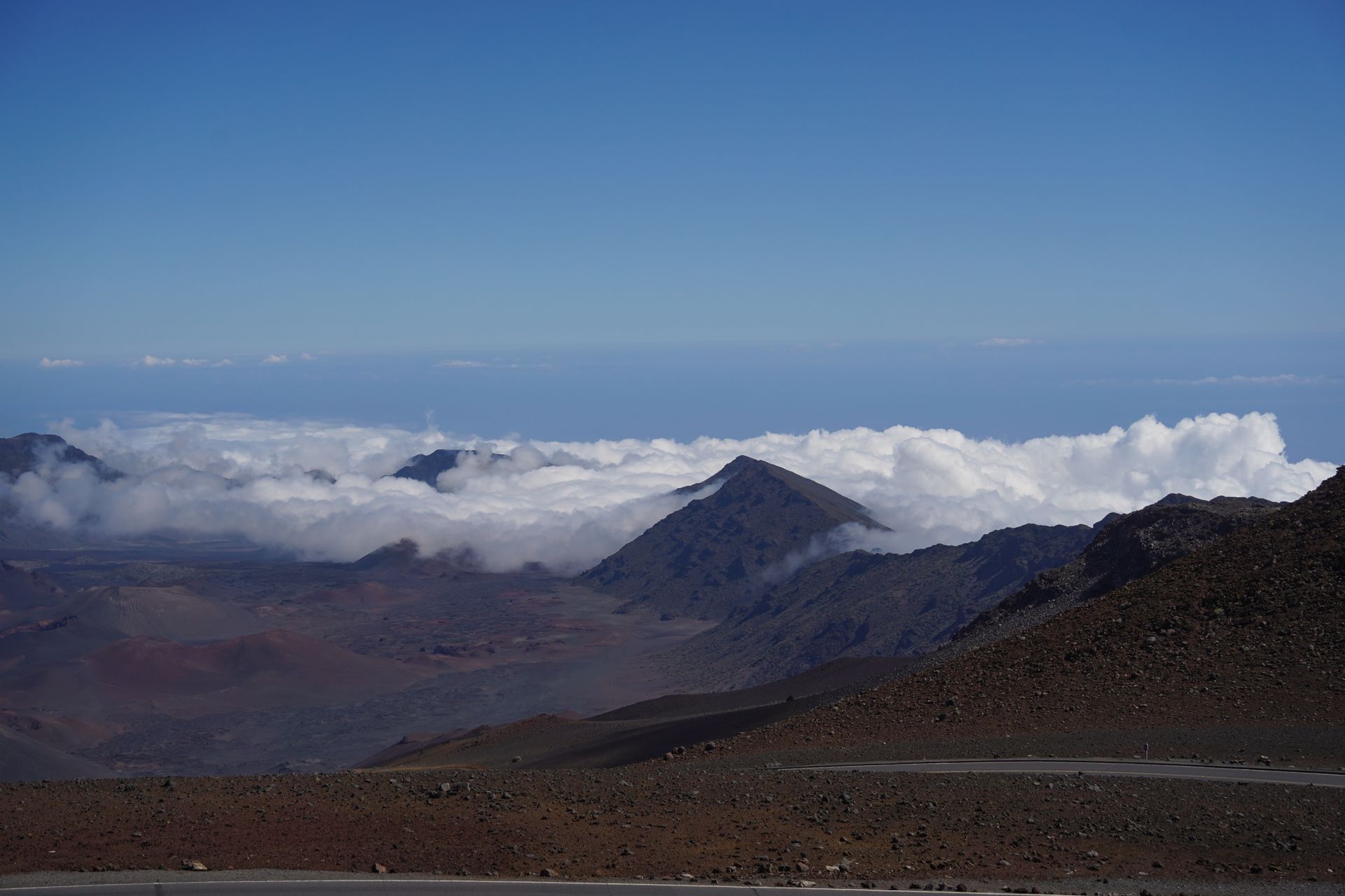 Looking down at clouds hugging reddish mountains, and a road going through the volcanic landscape
