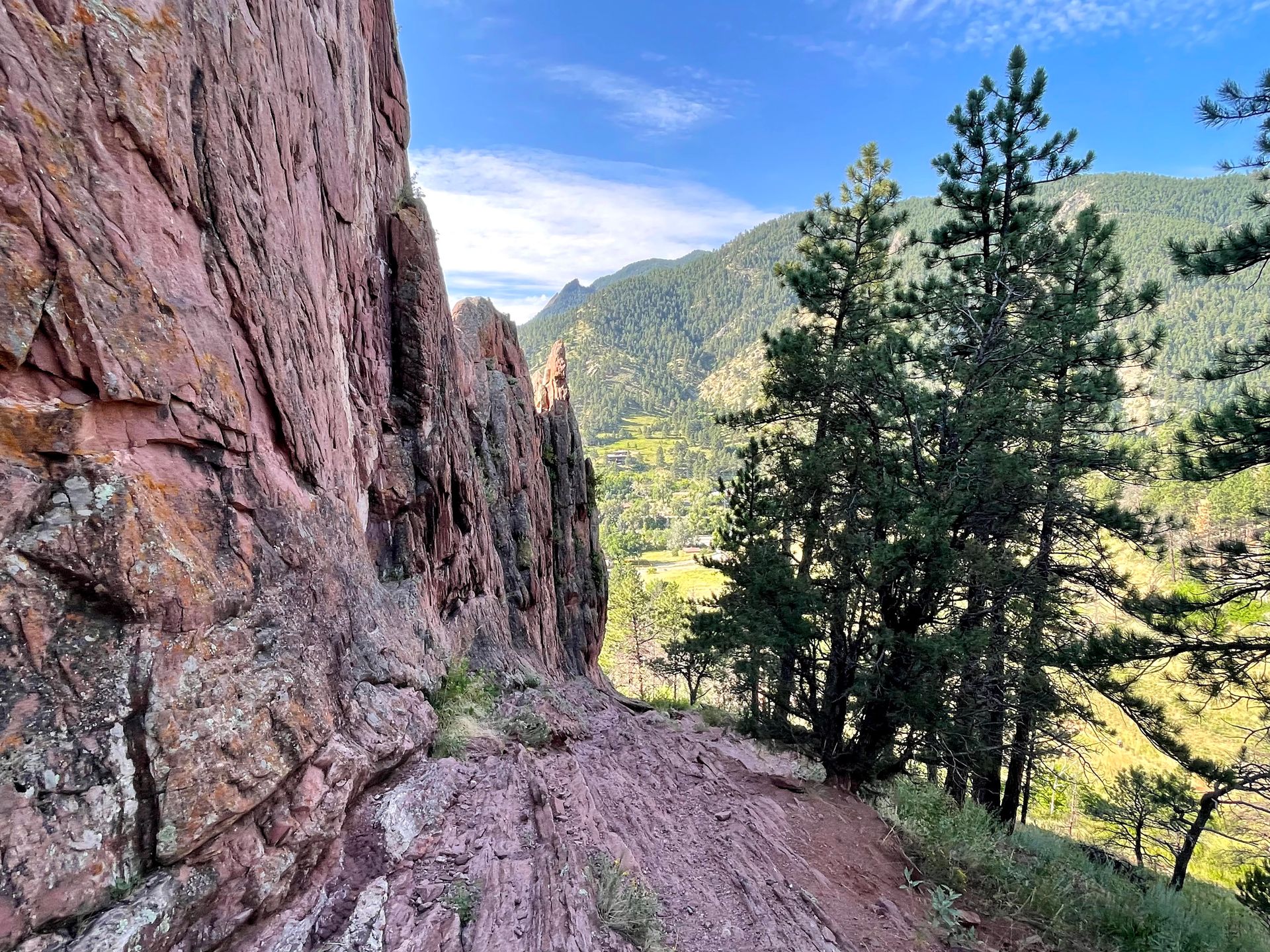 A red rock face with a trail next to it and green hills in the distance.