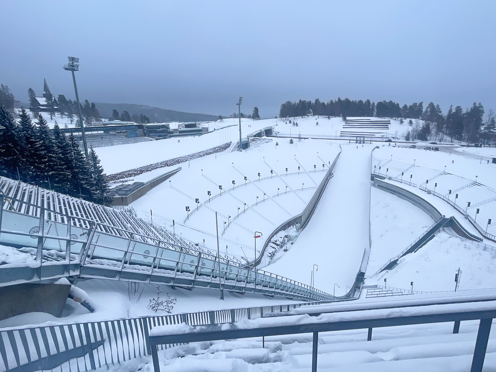 Looking down at a huge ski jump, surrounded by bleacher seats, covered in snow
