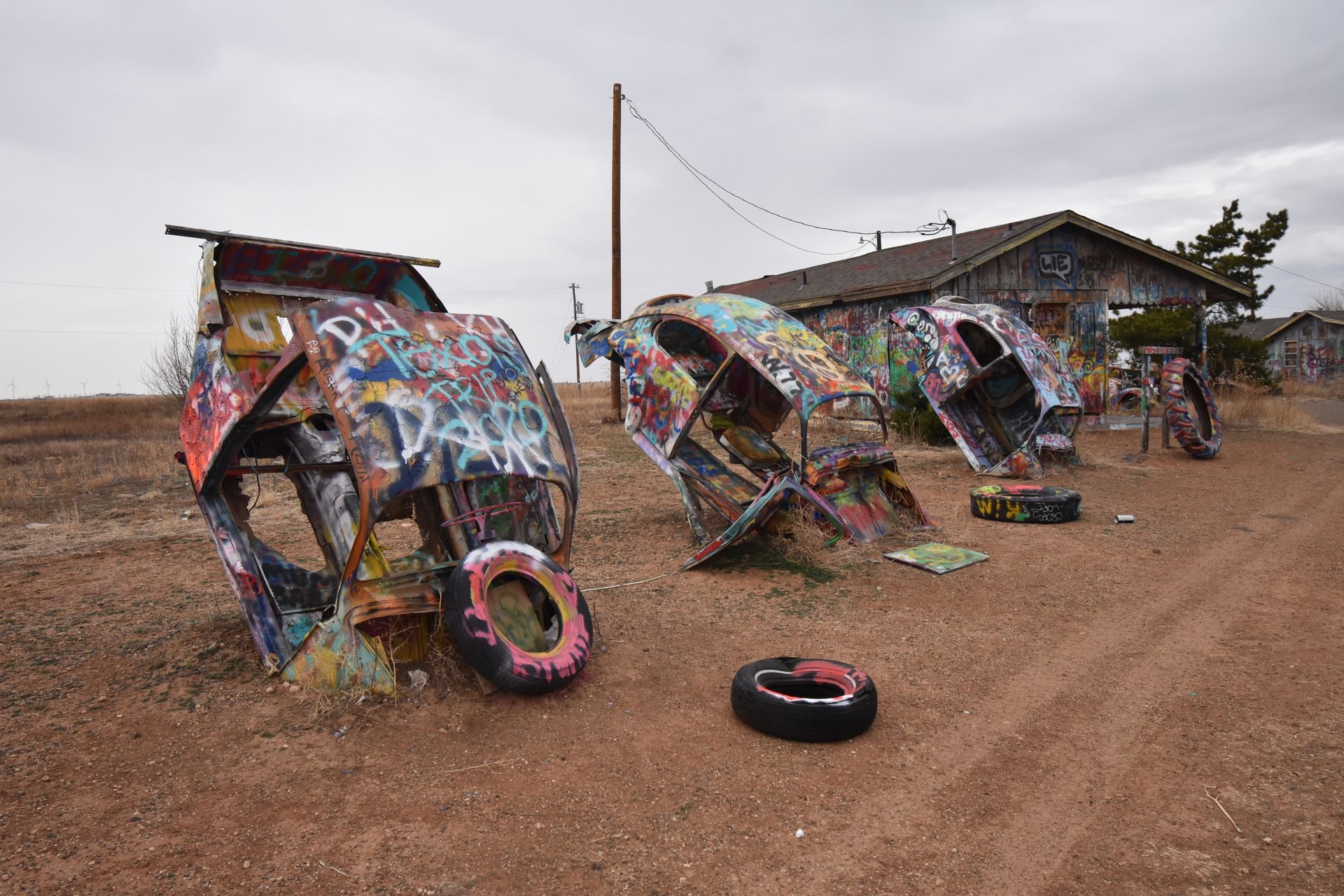 A row of three volkwagen beetles sticking up out of the ground covered in spray paint. There is a spray painted building in the background.