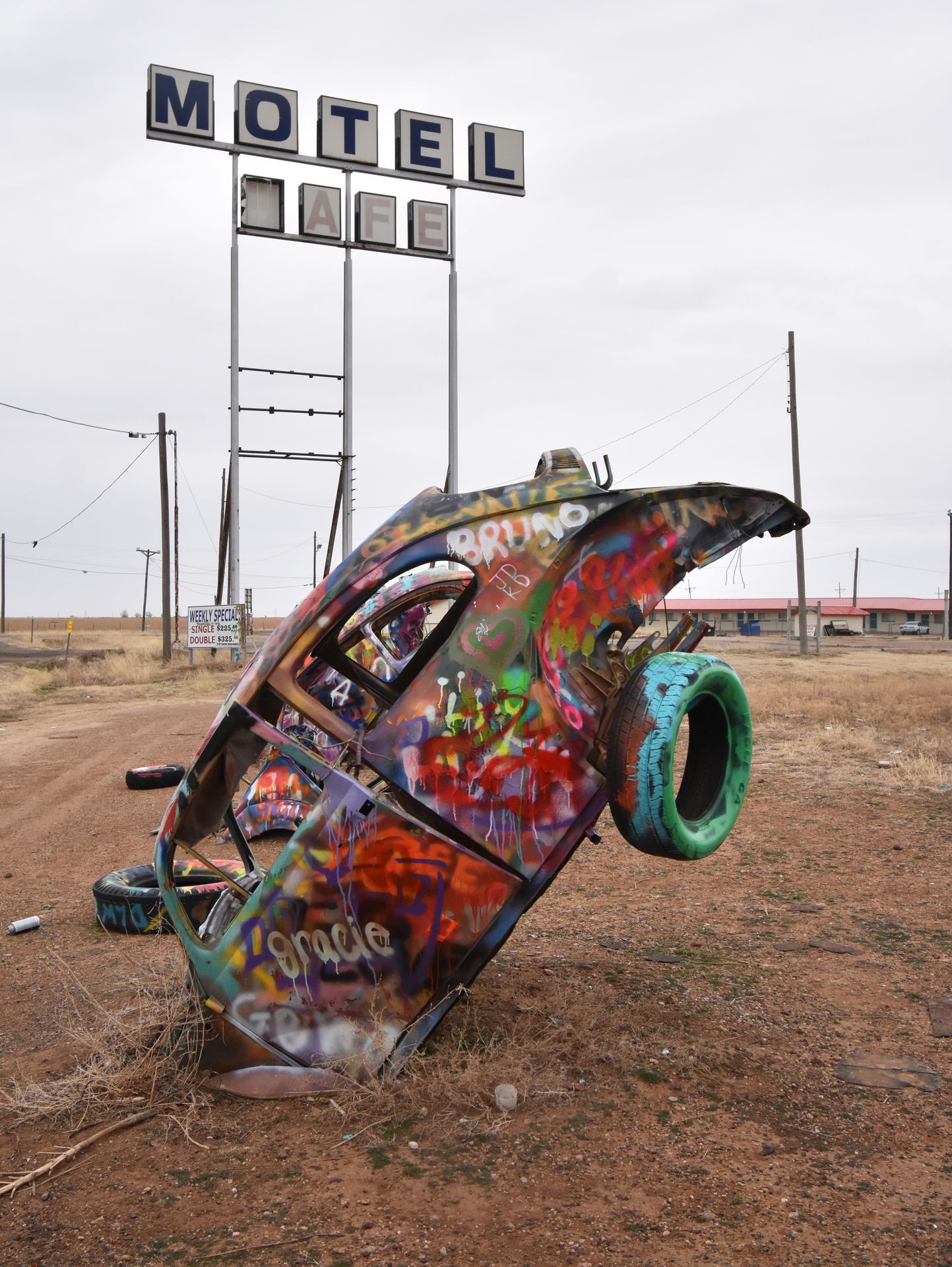 A volkwagen beetle sticking out of the ground covered in spray paint. There is a motel sign in the background.
