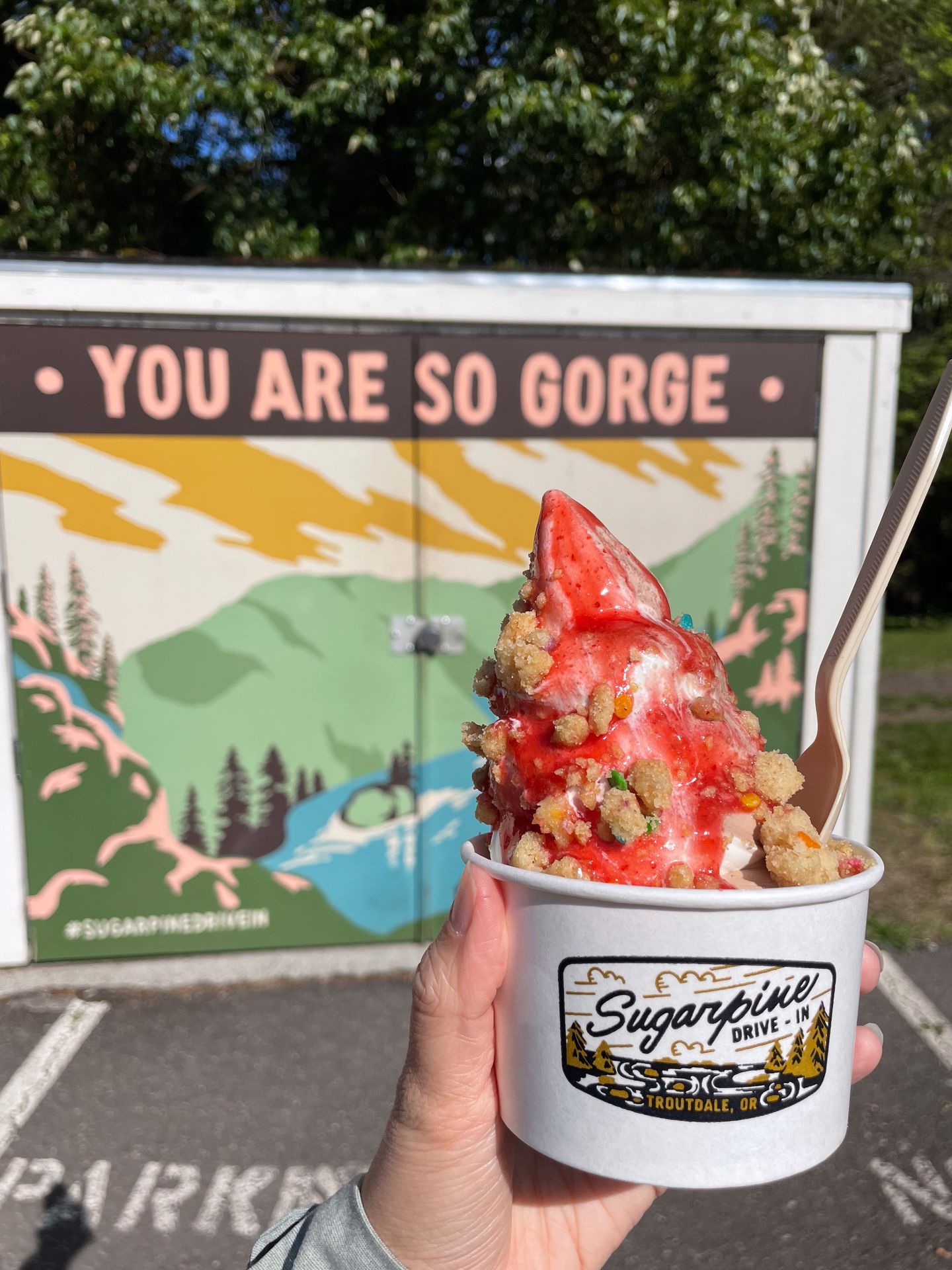 Holding up ice cream topped with red sauce and bits of cake in front of a mural that reads 'You are so Gorge'