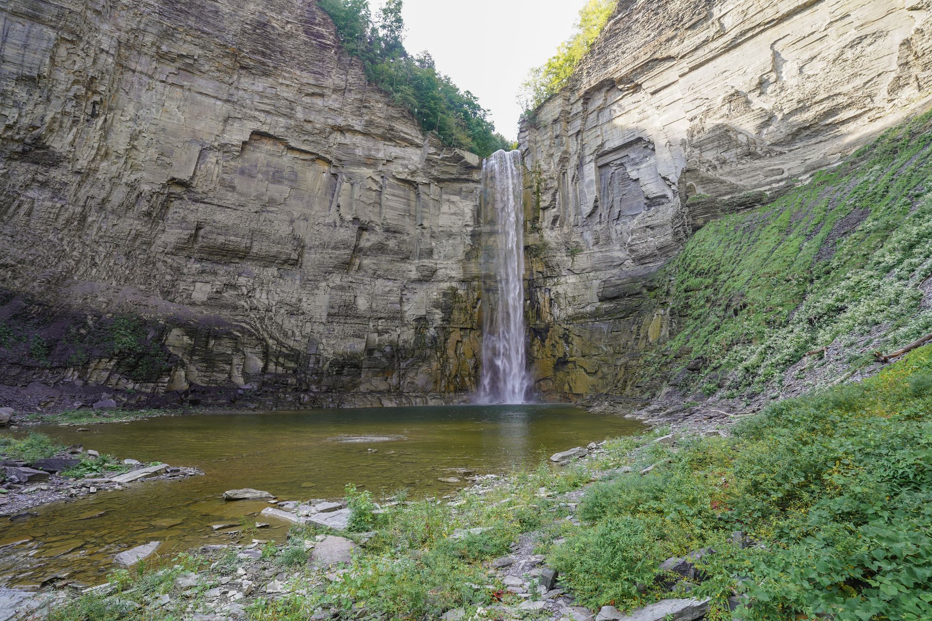 A tall waterfall falling down a tall rock wall and into a pool of water.